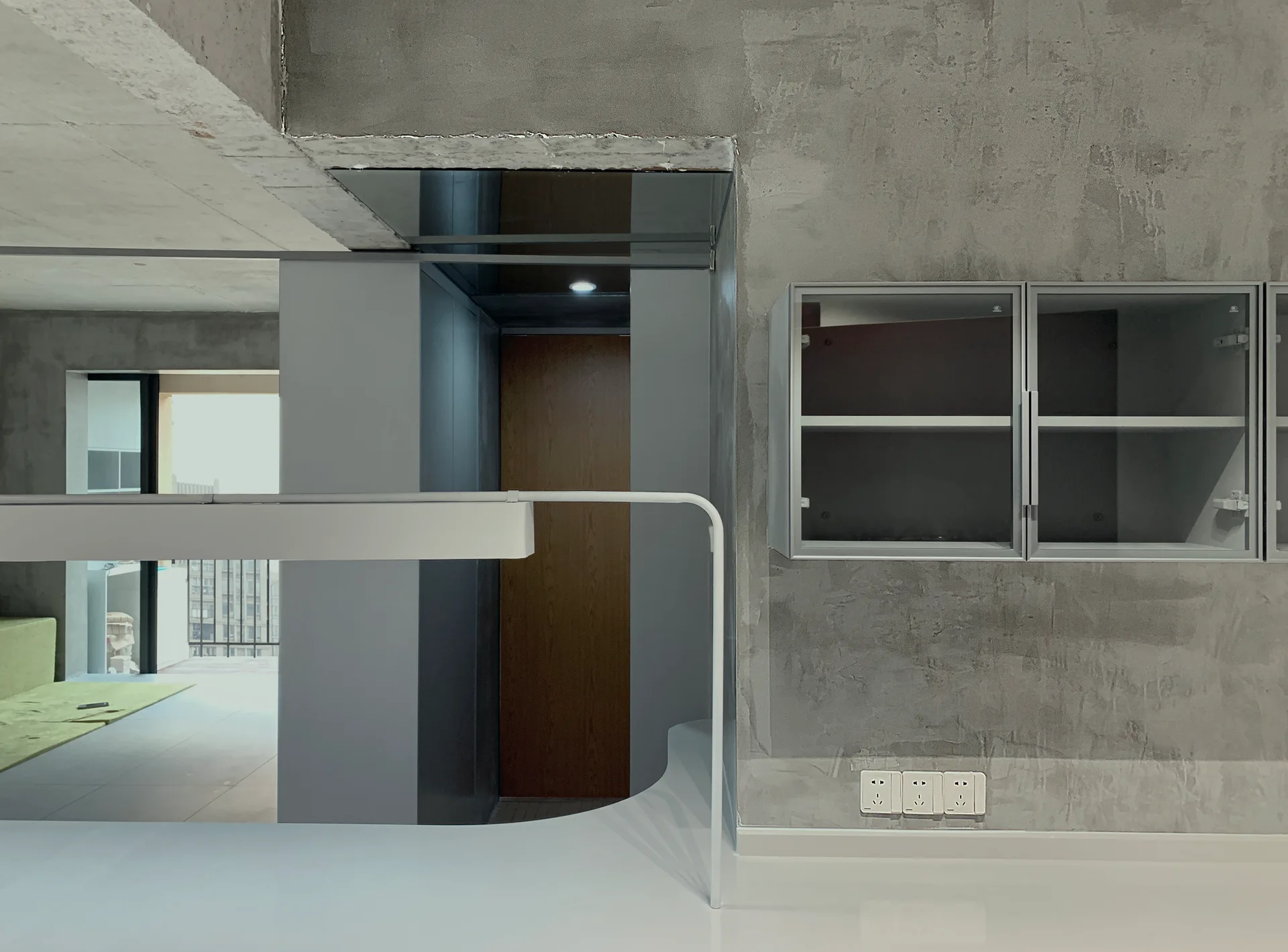 A minimalist interior featuring raw grey concrete walls, a silver metal wall cabinet with glass doors, and a sleek white modern desk fixture.