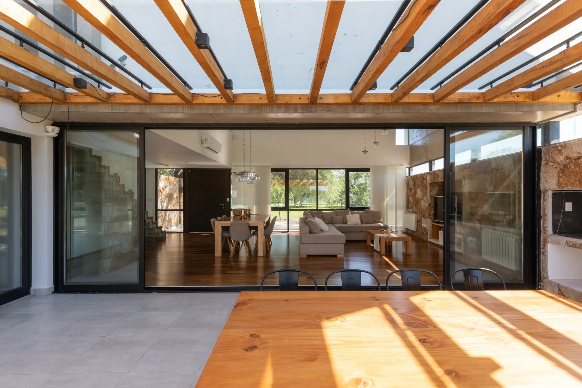 A view from a covered patio with wooden rafters looking through wide sliding glass doors into a modern open-plan living and dining room.