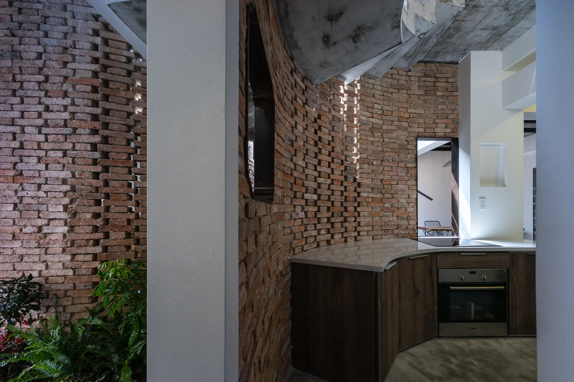 A modern kitchen featuring curved wood-toned cabinets, a built-in oven, and a dramatic curved red brick wall below a concrete spiral staircase.