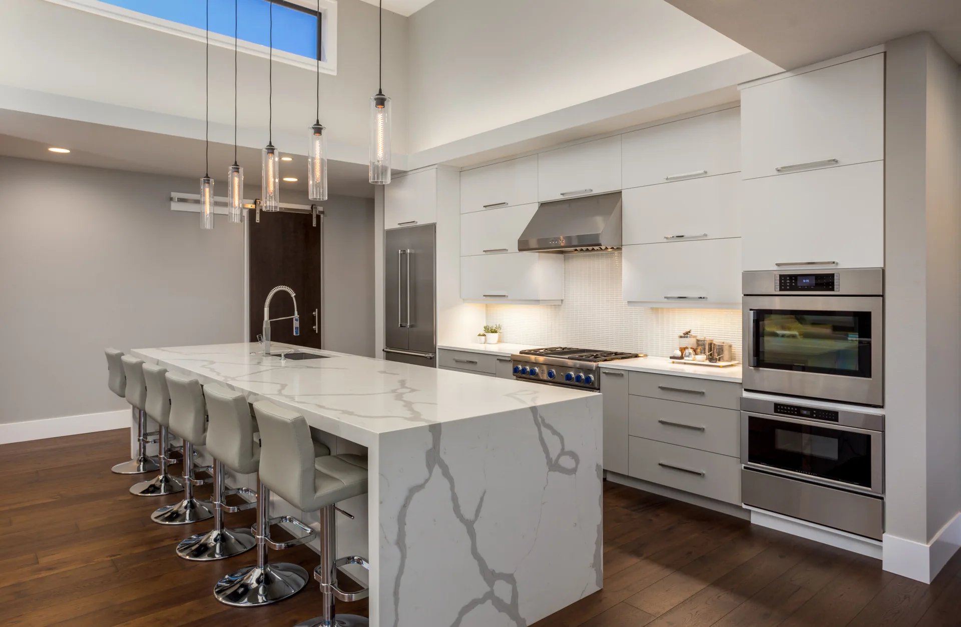 A modern kitchen featuring a large veined stone waterfall island with grey barstools, flat-panel white cabinets, and stainless steel appliances.