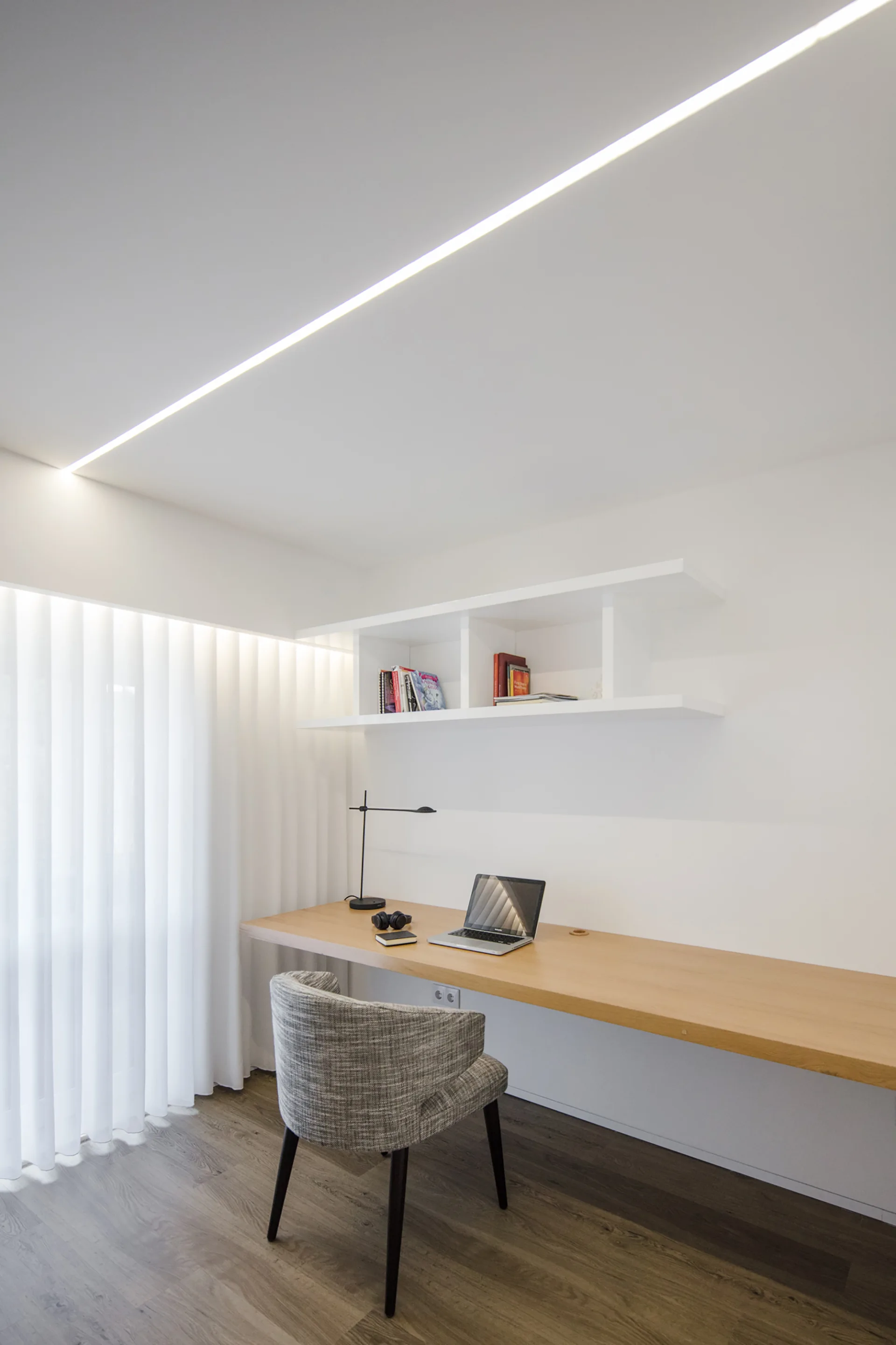 A minimalist home office featuring a long wood floating desk, grey upholstered chair, white shelving, and a recessed diagonal ceiling light.
