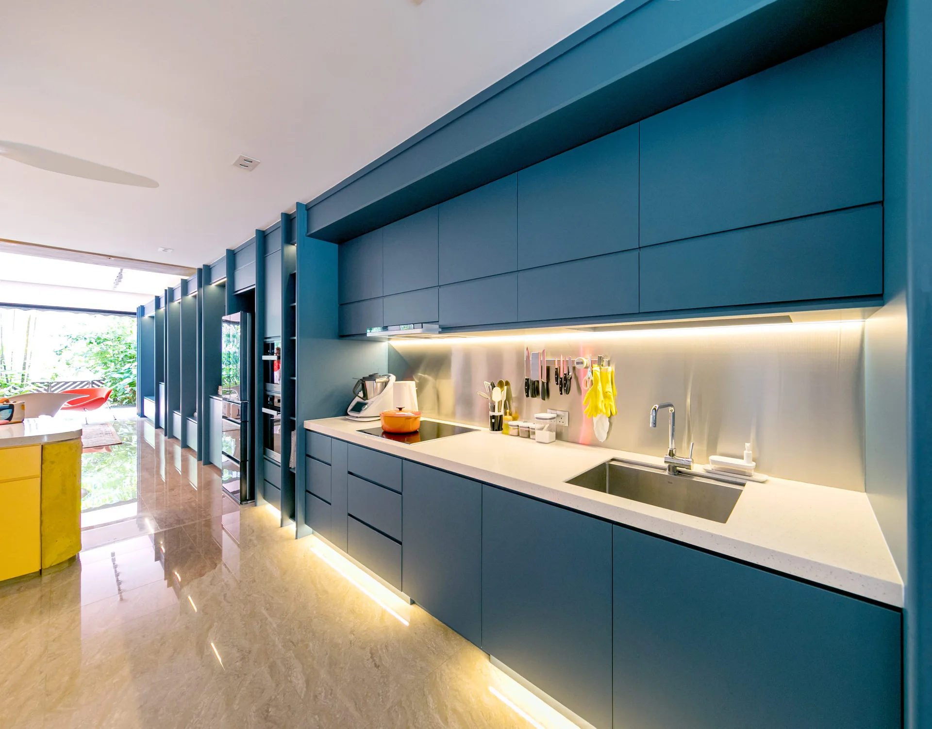 A modern kitchen features matte teal cabinets, a metallic backsplash, a stainless steel sink, and a yellow island base on a glossy marble floor.