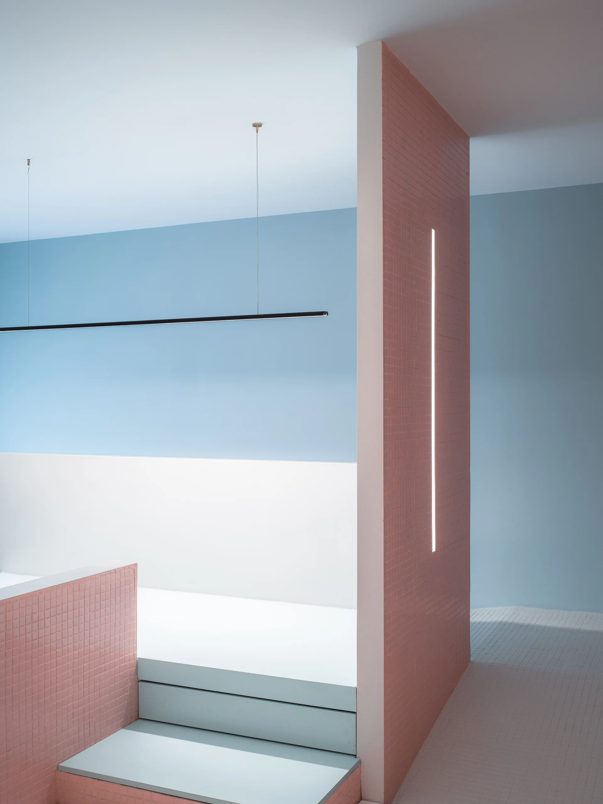 A minimalist interior space featuring a pink tiled partition wall with an integrated vertical light strip next to light blue steps and walls.