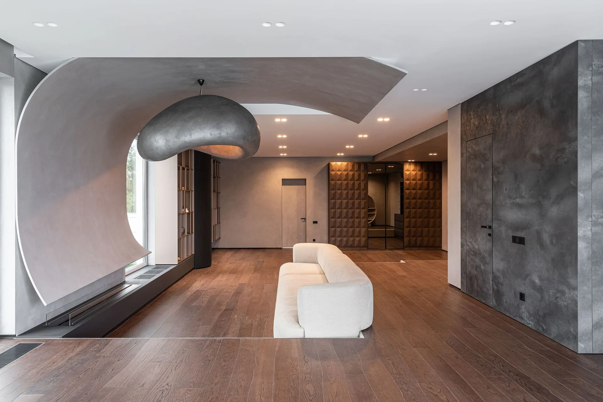 A minimalist living room features a cream sofa on dark wood floors beneath a large sculptural pendant light and a curved concrete wall feature.