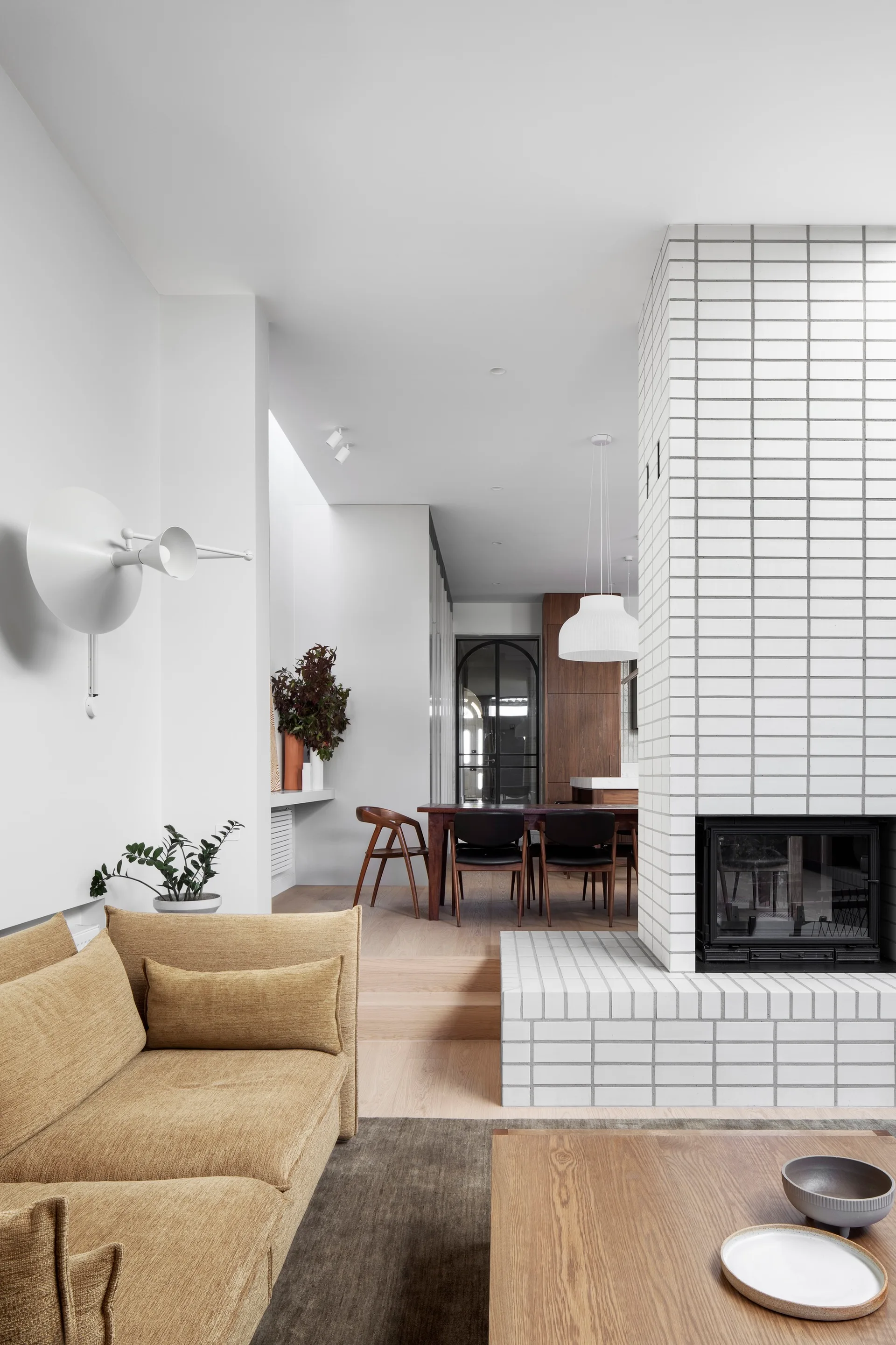 A split-level living room features a mustard sofa and a white grid-tiled fireplace column next to steps leading up to a dining area.