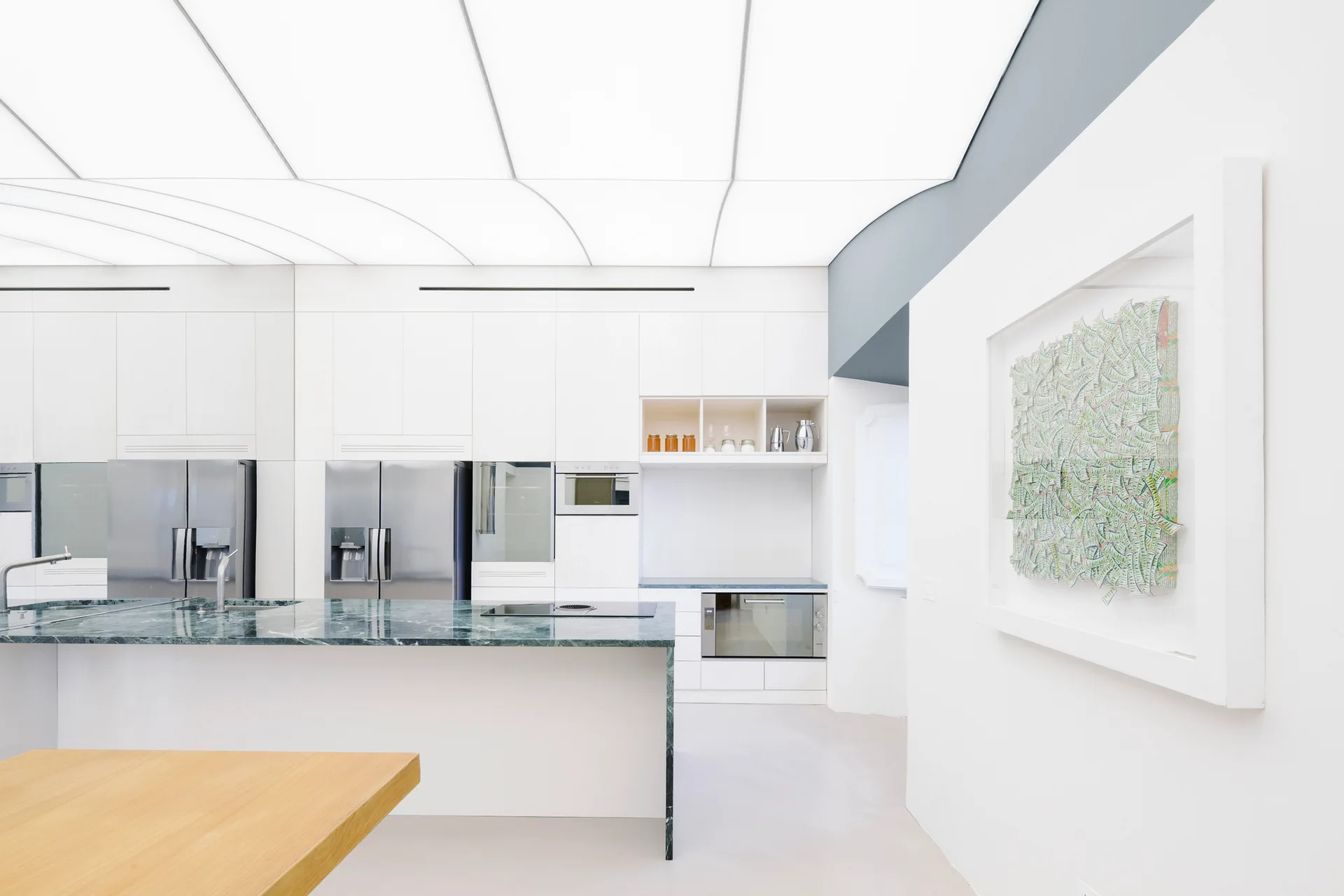 A minimalist white kitchen features a large green marble island, stainless steel appliances, and a glowing backlit grid ceiling.
