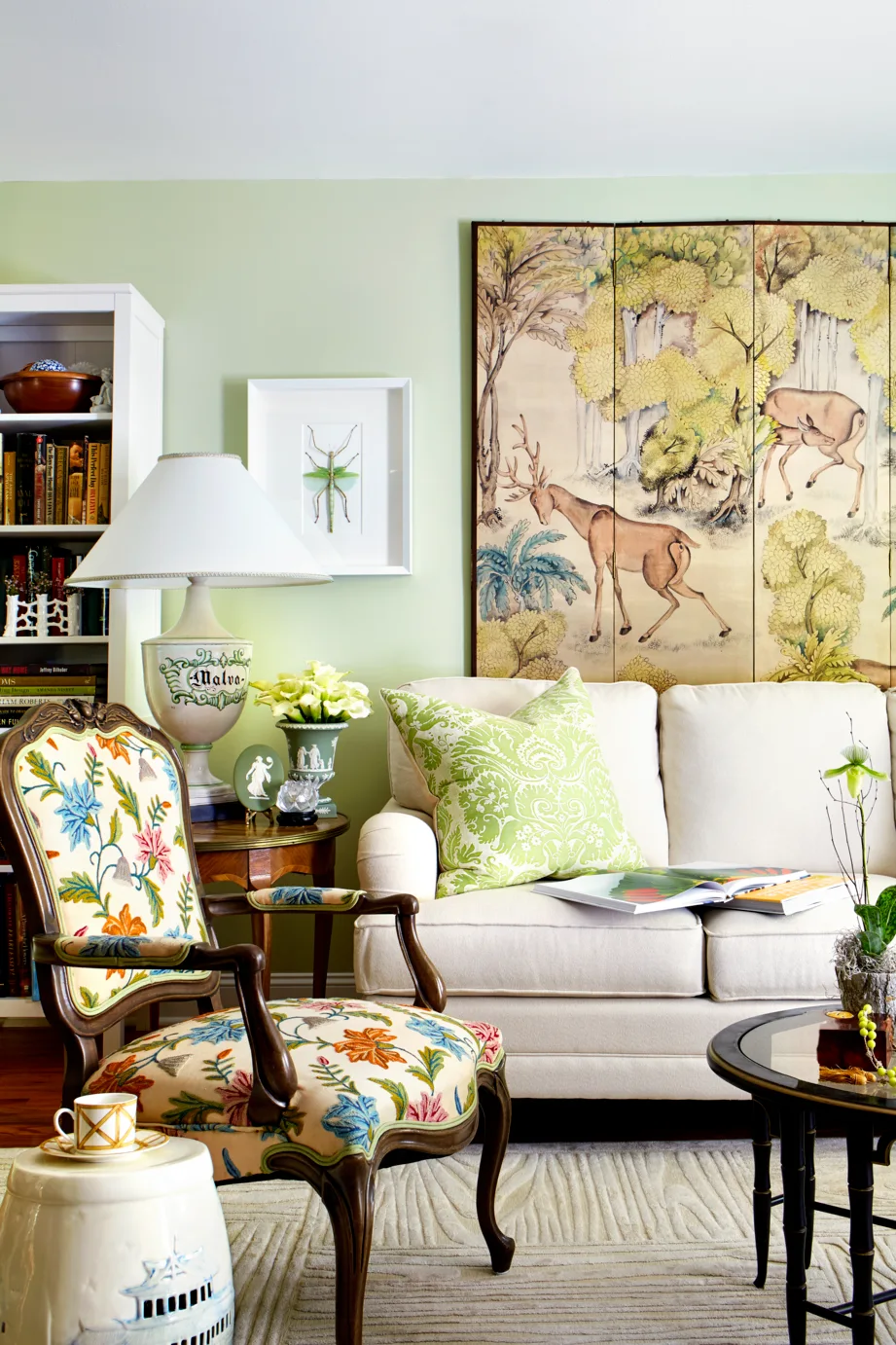 A living room featuring a cream sofa, a floral embroidered armchair, and a large painted folding screen with deer imagery mounted on a pale green wall.