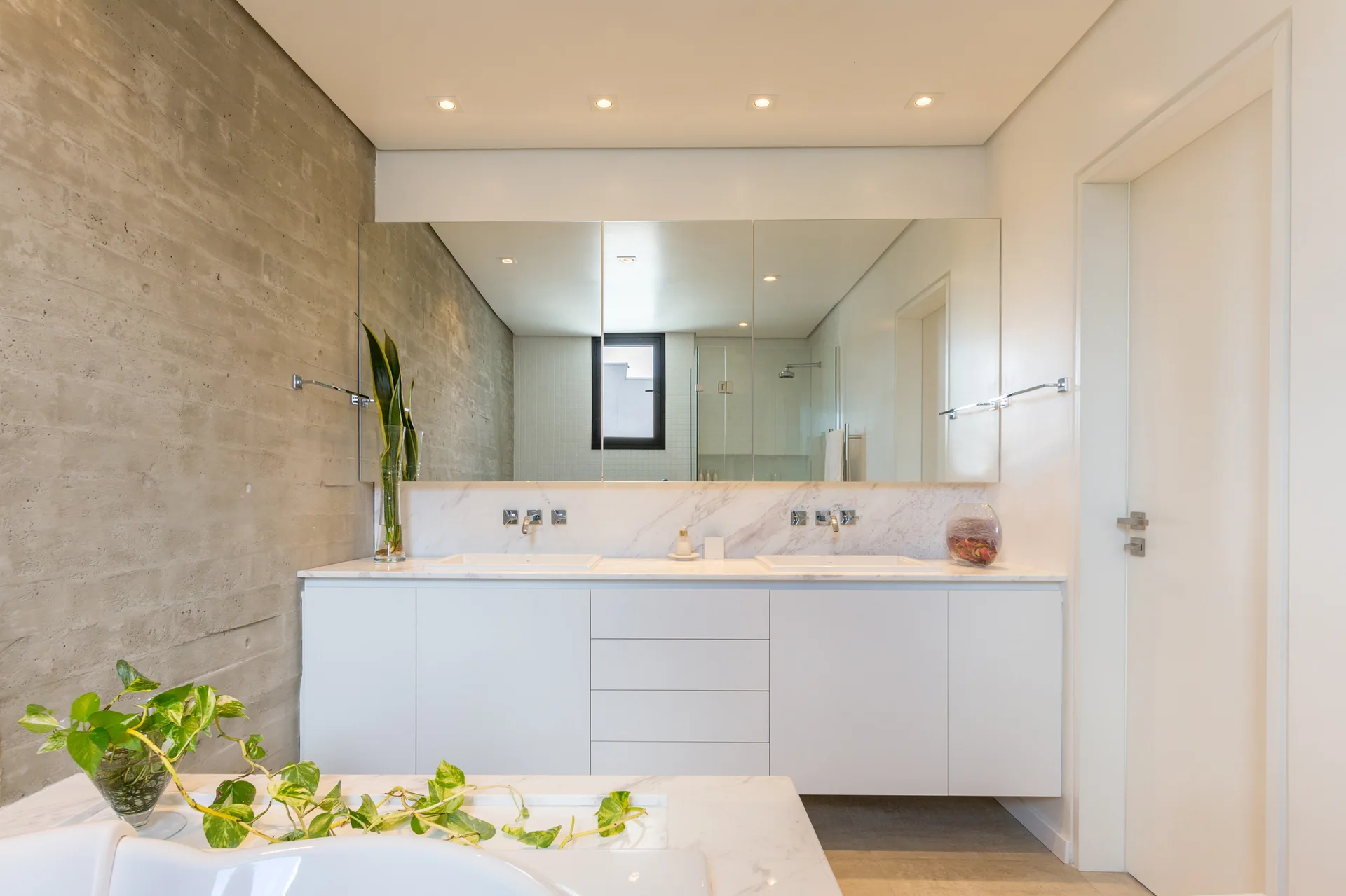 A modern bathroom features a floating white double vanity with a marble countertop, set against a textured raw concrete accent wall and a large mirror.