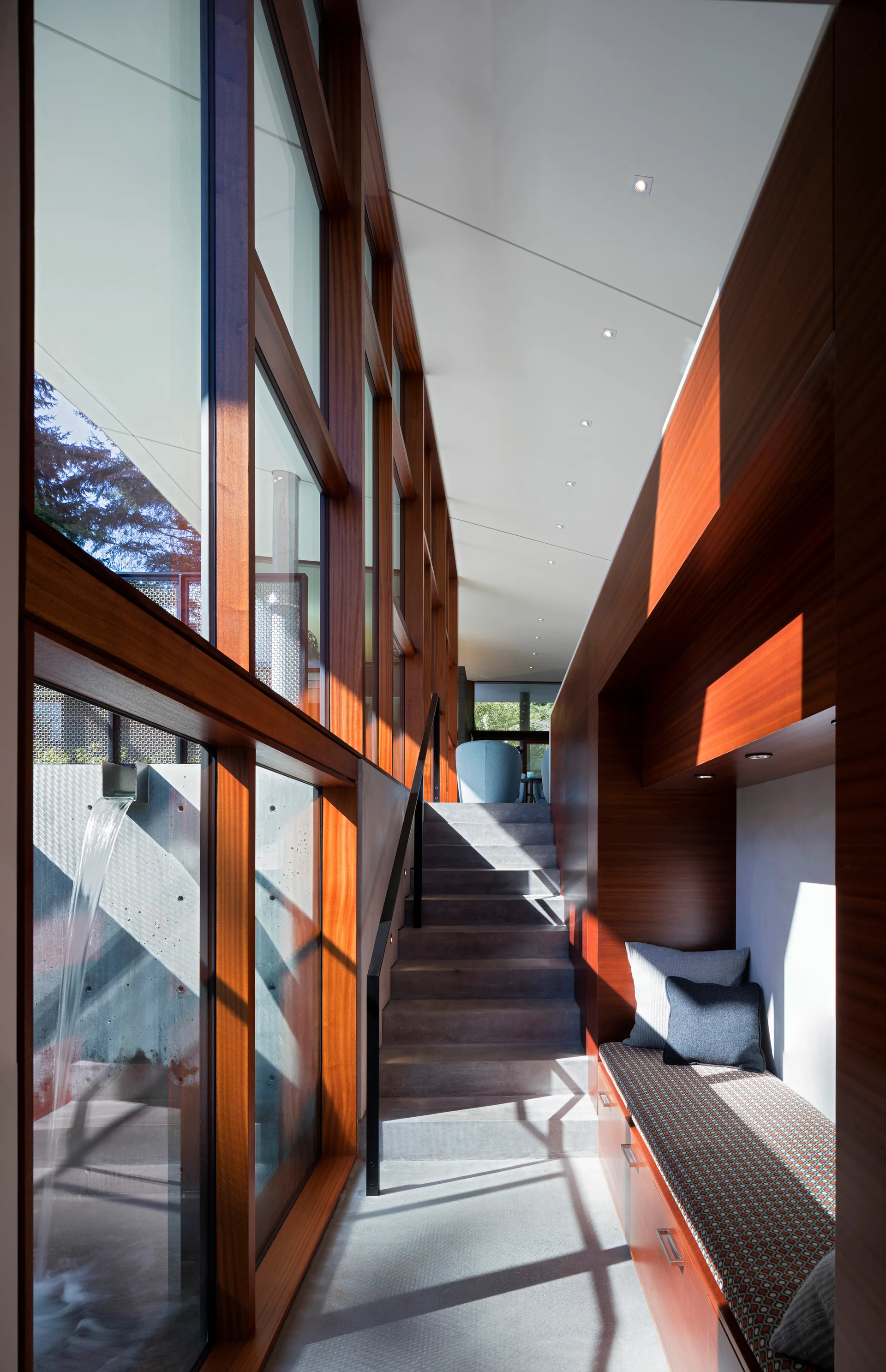 A modern staircase features concrete steps, warm wood wall paneling with a built-in bench, and floor-to-ceiling wooden framed windows.