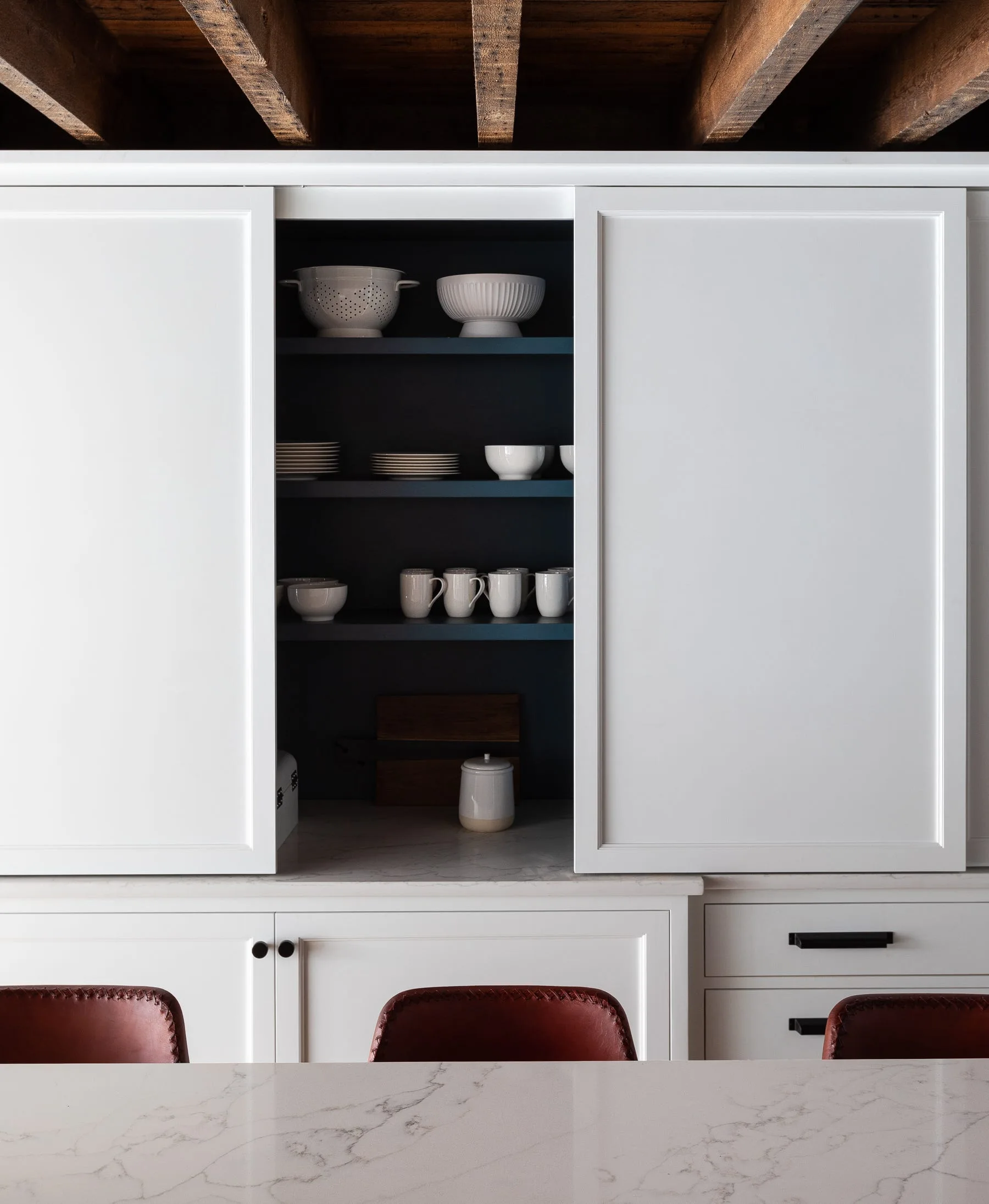 White sliding cabinet doors open to reveal a dark grey pantry shelf stocked with white dishware above a marble countertop and below exposed wooden beams.