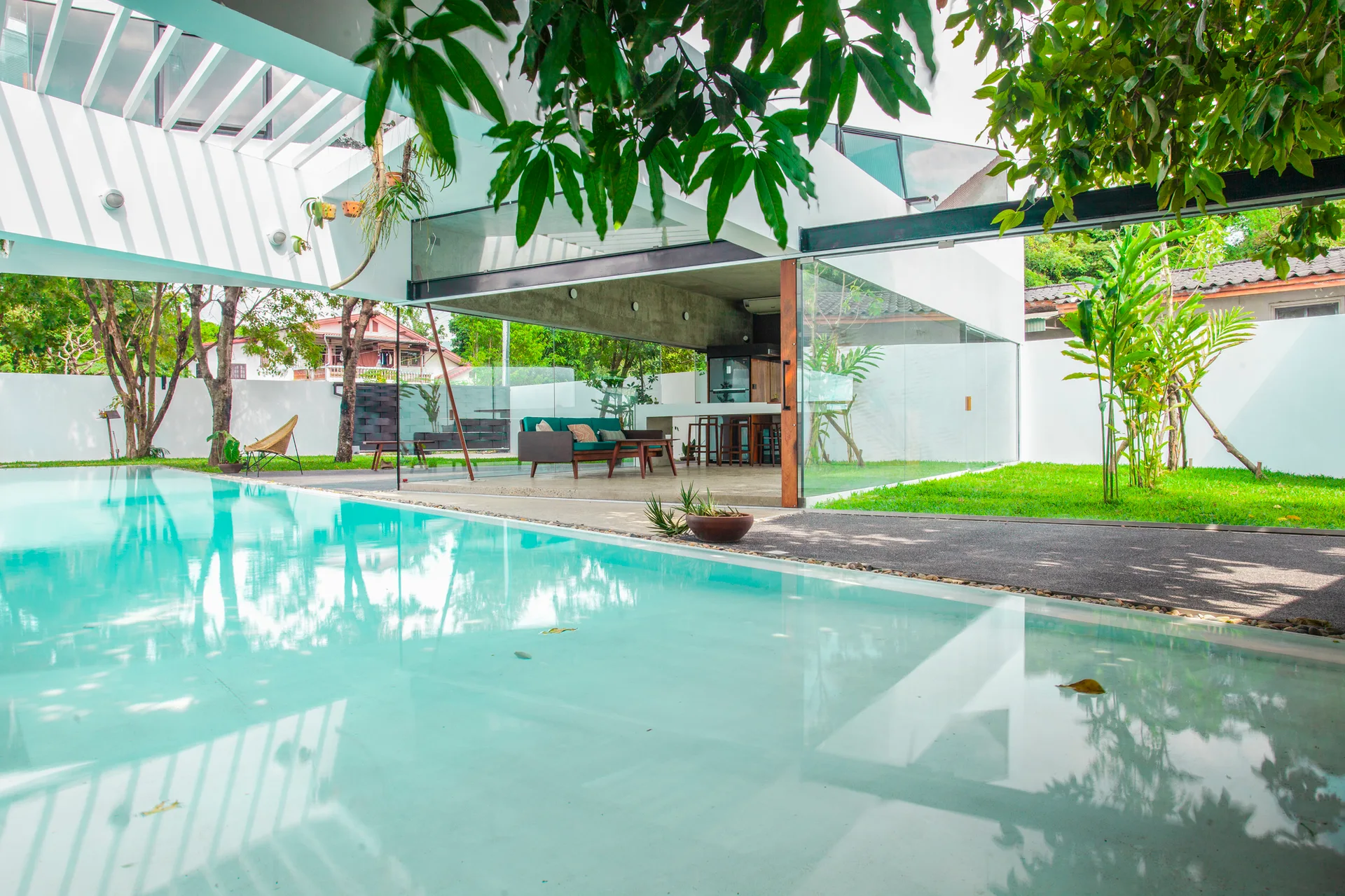 A modern minimalist glass pavilion with a green sofa overlooks a clear swimming pool flanked by a white slatted pergola and green lawn.
