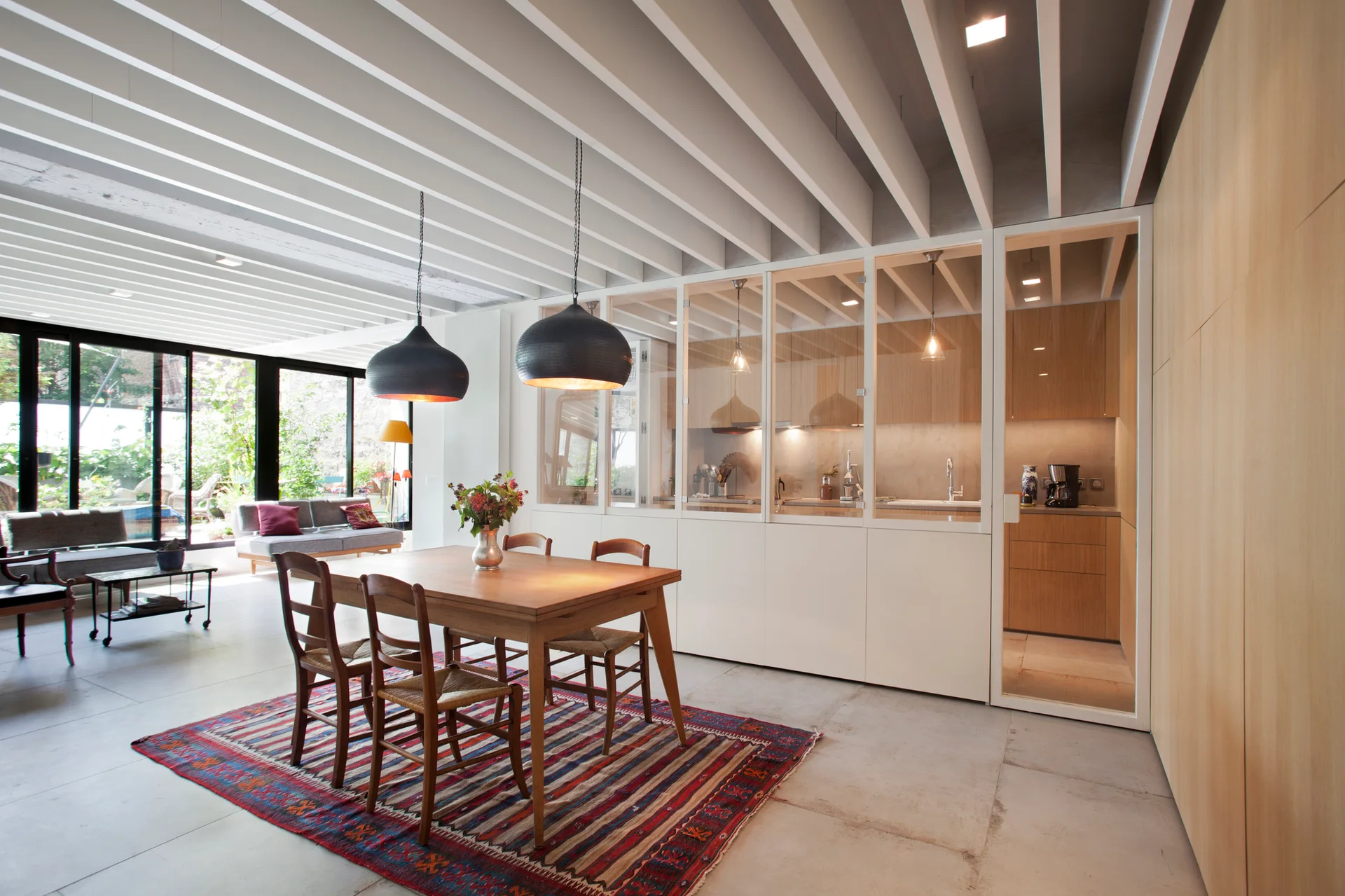 A wooden dining table and chairs sit on a striped rug beneath dark pendant lights in front of a glass-walled kitchen partition.