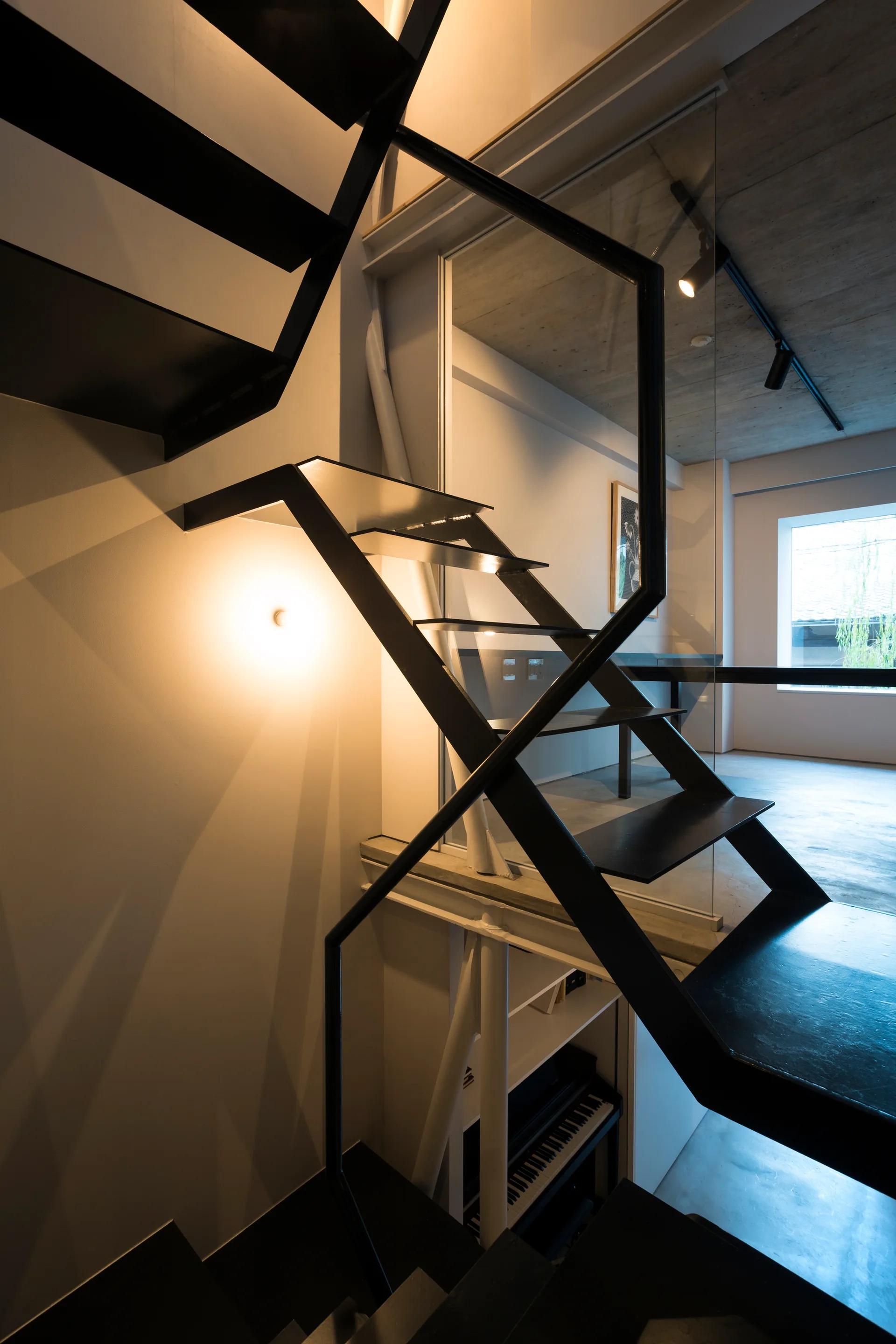 An industrial style black steel open riser staircase stands next to a glass partition wall with a piano visible underneath.
