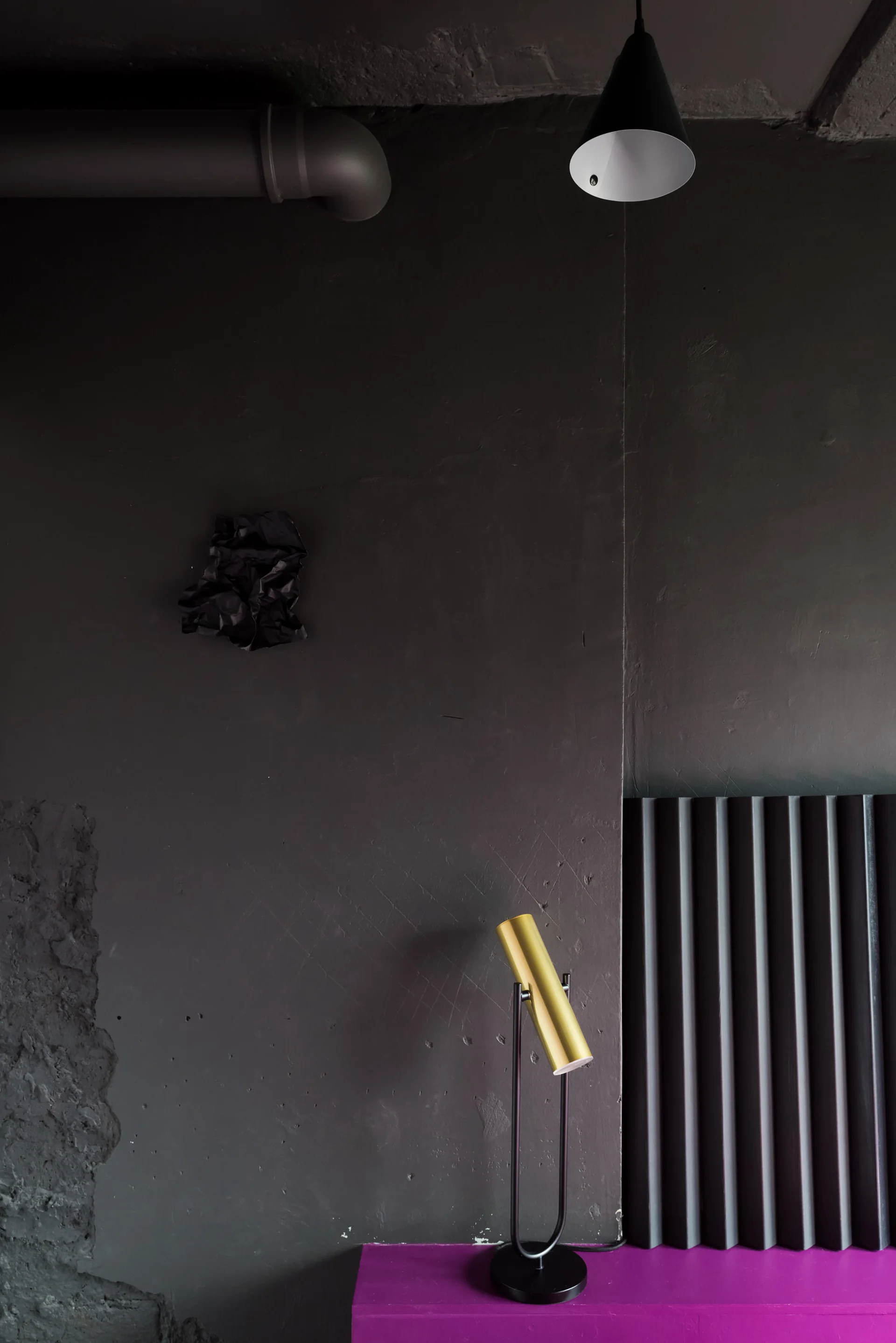 A cylindrical gold lamp sits on a bright magenta console against a matte charcoal wall featuring a fluted black panel and an exposed ceiling pipe.