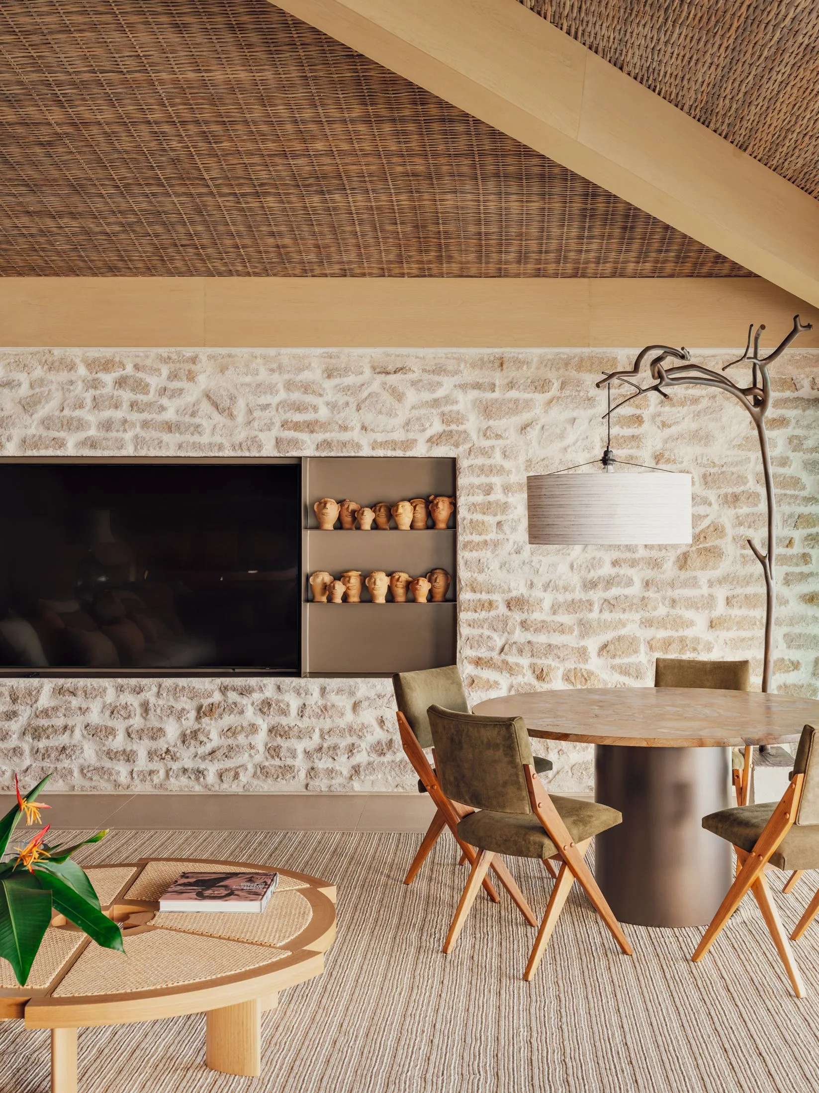 An organic modern dining room features a rugged stone accent wall with built-in niches, olive chairs, and a distinctive woven rattan ceiling.