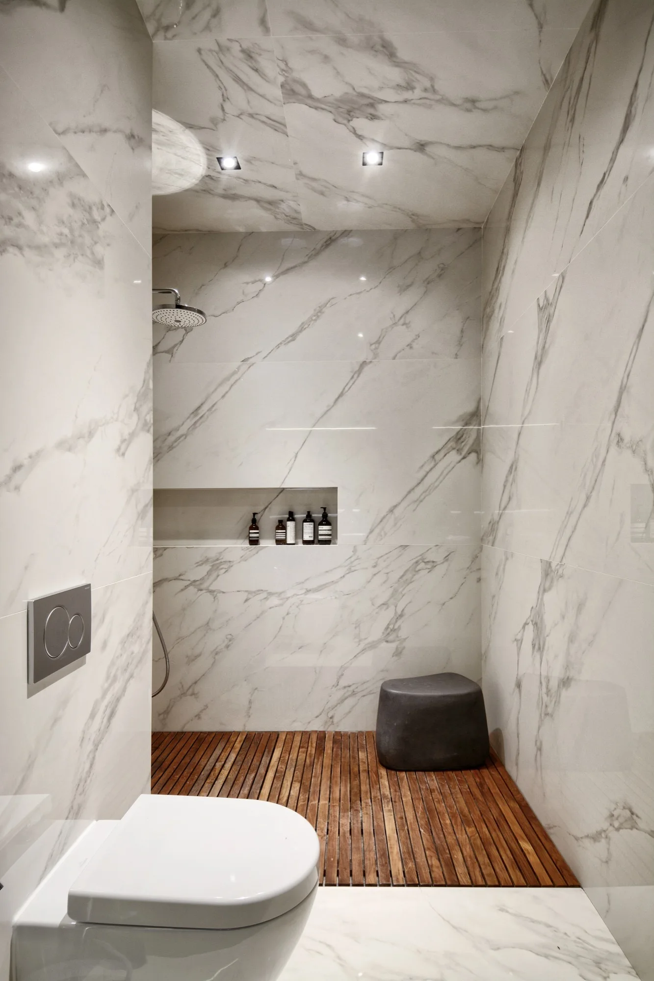 A modern bathroom clad in veined white marble featuring a wood slat shower floor, wall-hung toilet, and a recessed wall niche.