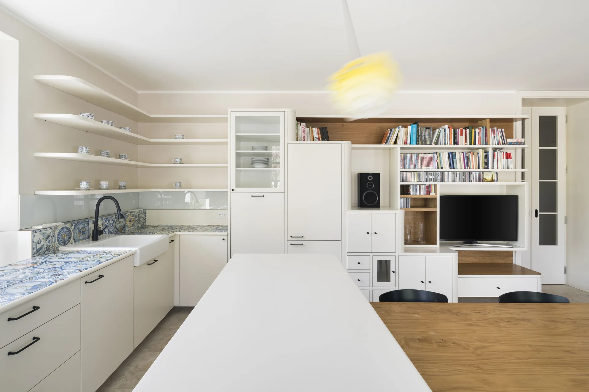 A modern white kitchen features blue patterned tile countertops, a farmhouse sink, and a large built-in shelving unit with books and a television.