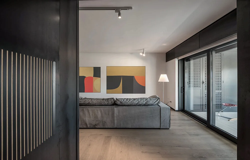 A minimalist living room features a low-profile gray sofa, two abstract paintings on a white wall, and sliding glass patio doors.