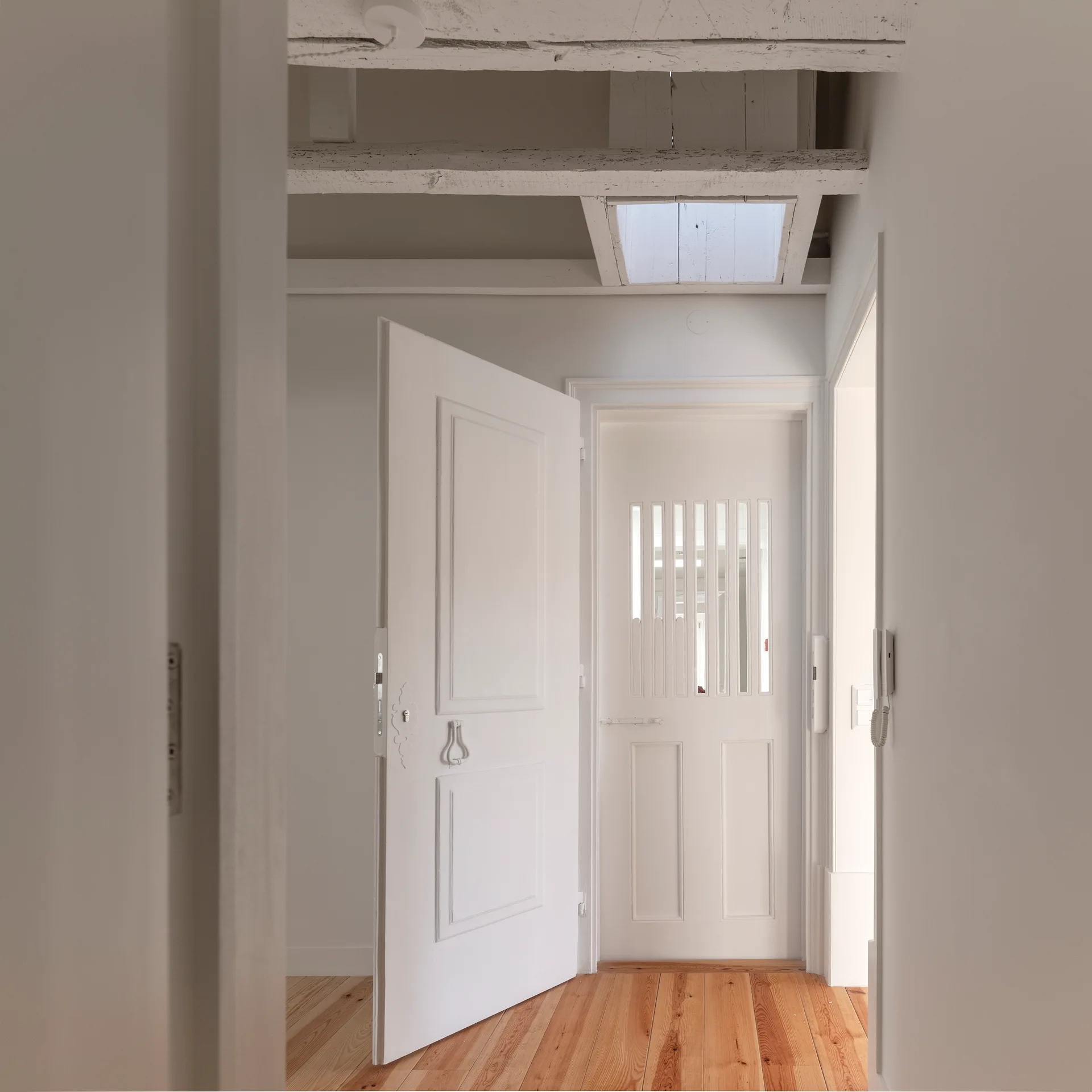 A minimalist white hallway features exposed ceiling beams, a small skylight, warm wood flooring, and paneled white doors.