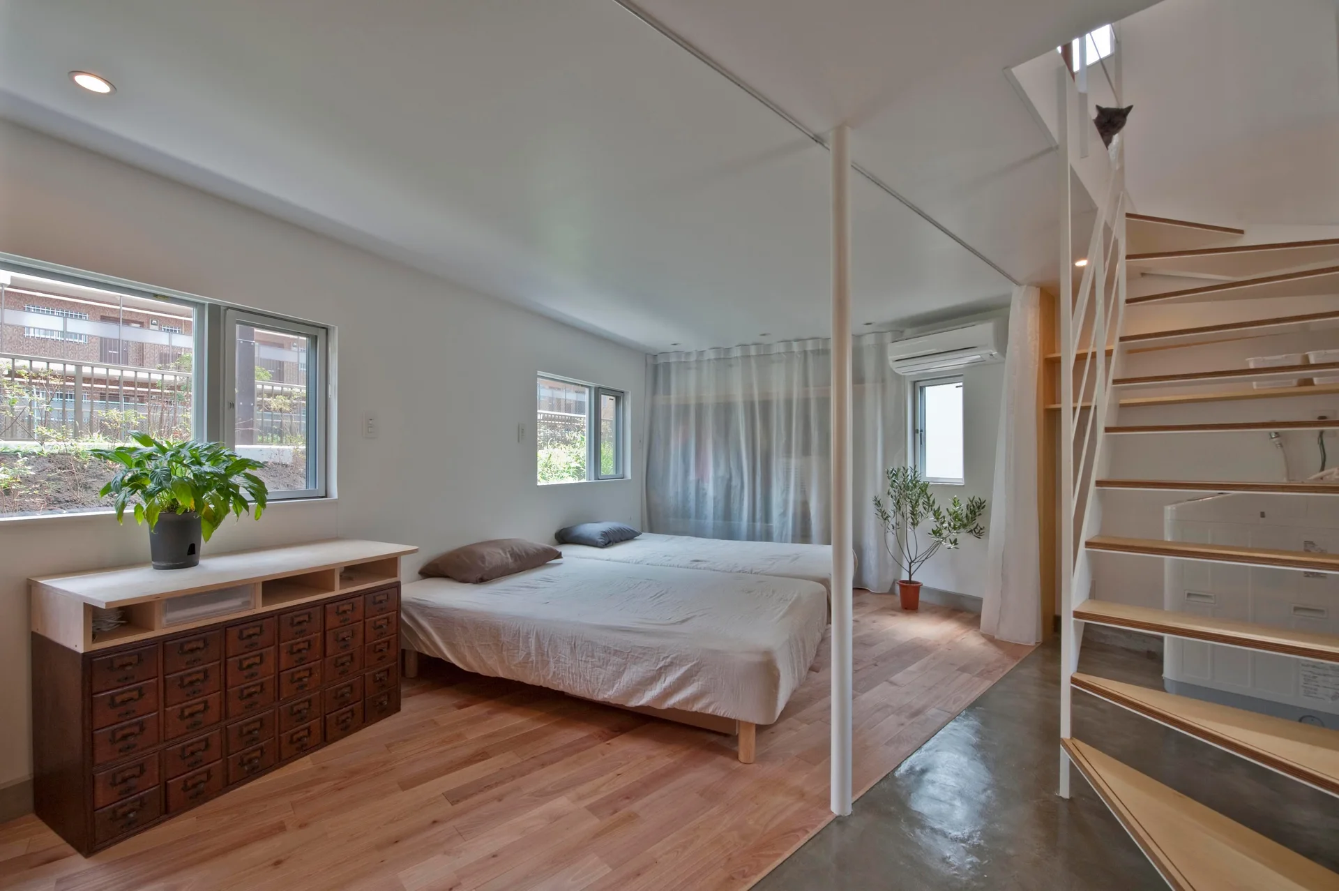 A minimalist bedroom featuring two low twin platform beds, a dark wood apothecary chest, and a white spiral staircase on mixed wood and concrete floors.