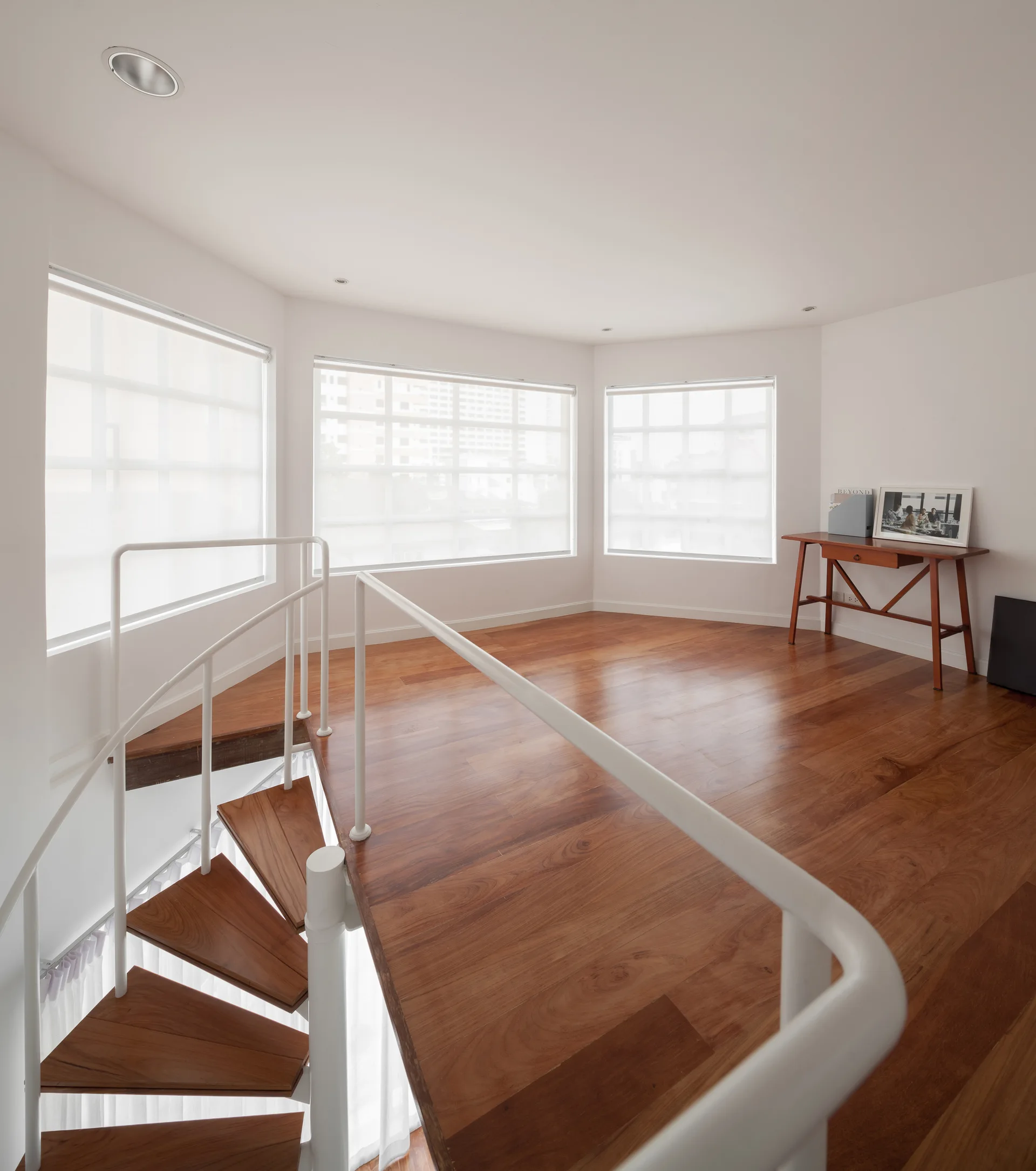 A bright interior landing features a white spiral staircase with wood treads, polished hardwood floors, and three windows with white roller shades.