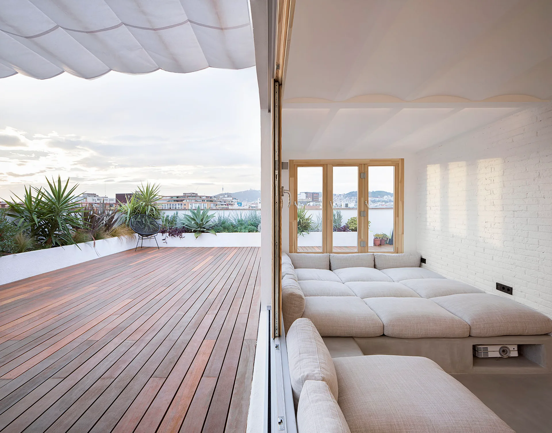 A wooden rooftop terrace with green plants next to an indoor lounge featuring a low built-in platform bed with beige cushions.
