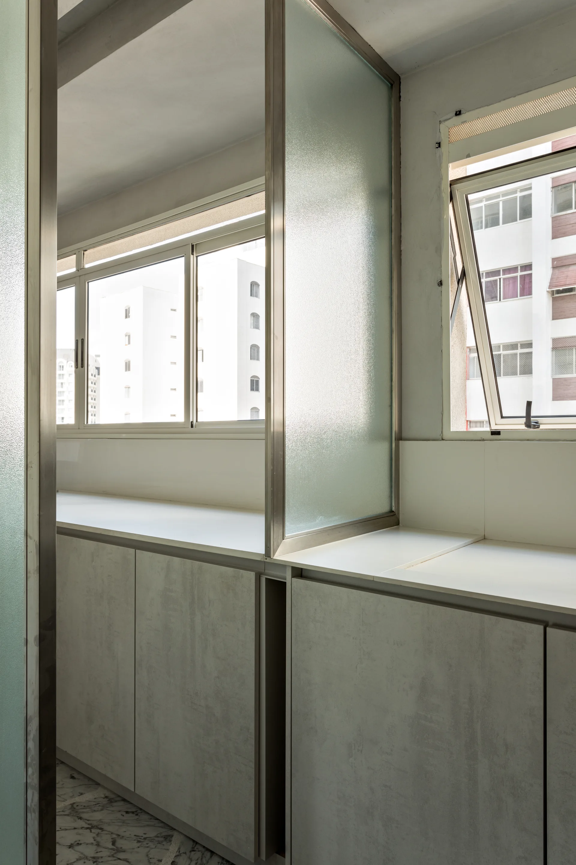 Modern grey textured base cabinets sit beneath a white countertop with a metal-framed frosted glass partition panel near bright windows.