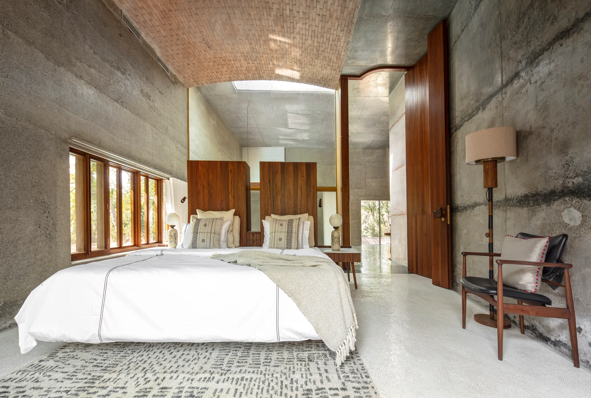 A modern bedroom features a vaulted brick ceiling, raw concrete walls, a bed with tall wooden headboard panels, and a patterned rug.