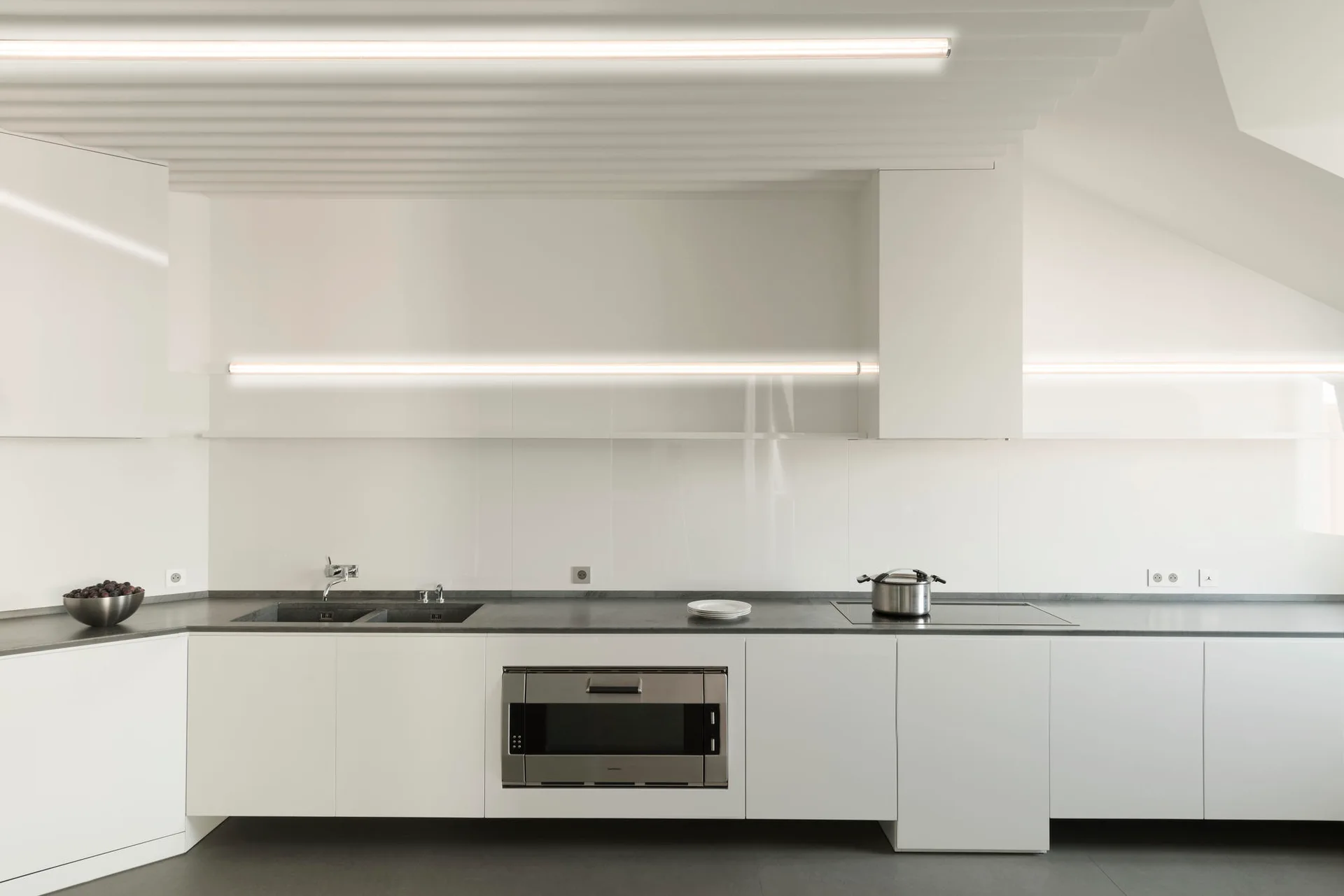 A minimalist white kitchen features flat-front cabinets, a dark grey countertop, integrated appliances, and long horizontal tube light fixtures.
