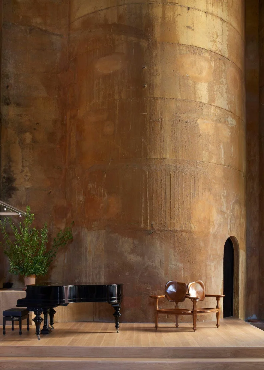 A glossy black grand piano and two wooden chairs sit on a light wood platform before a massive, textured rust-colored cylindrical wall.