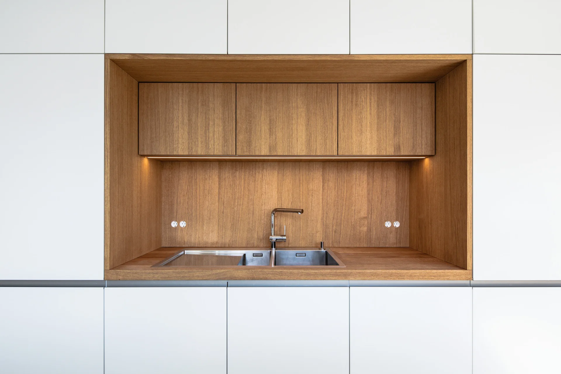 A stainless steel double sink set into a recessed wood-paneled niche within a seamless wall of flat-panel white kitchen cabinets.