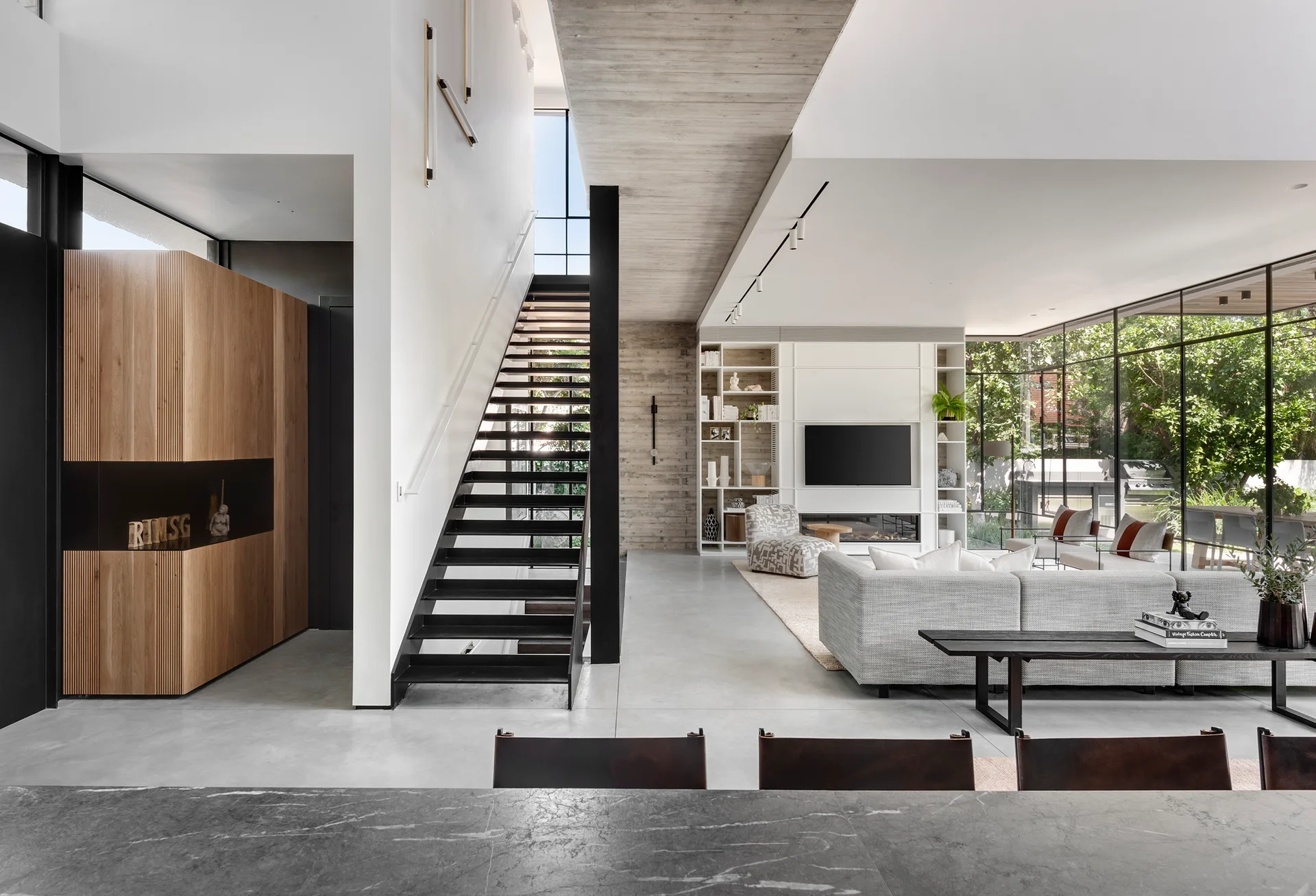 A modern open-plan living room featuring a black metal staircase, an exposed concrete ceiling beam, a grey sofa, and large windows.