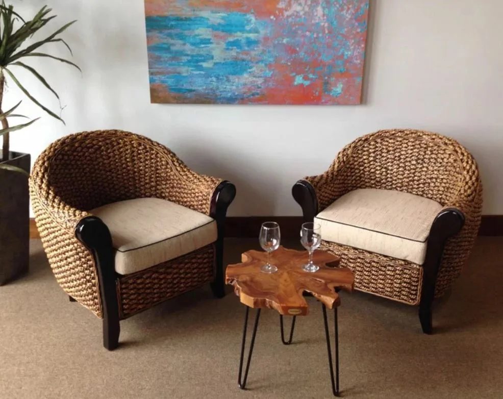 Two woven wicker armchairs with beige cushions flank a small live-edge wood side table with hairpin legs in front of an abstract wall painting.