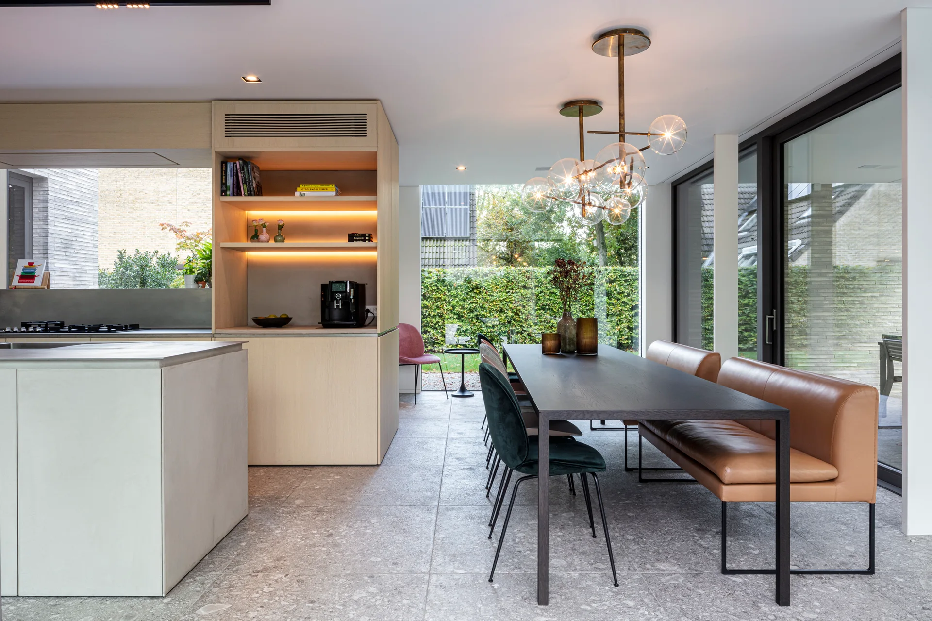 A modern open-plan dining space featuring a dark wood table paired with a tan leather banquette, grey stone floors, and a brass globe chandelier.