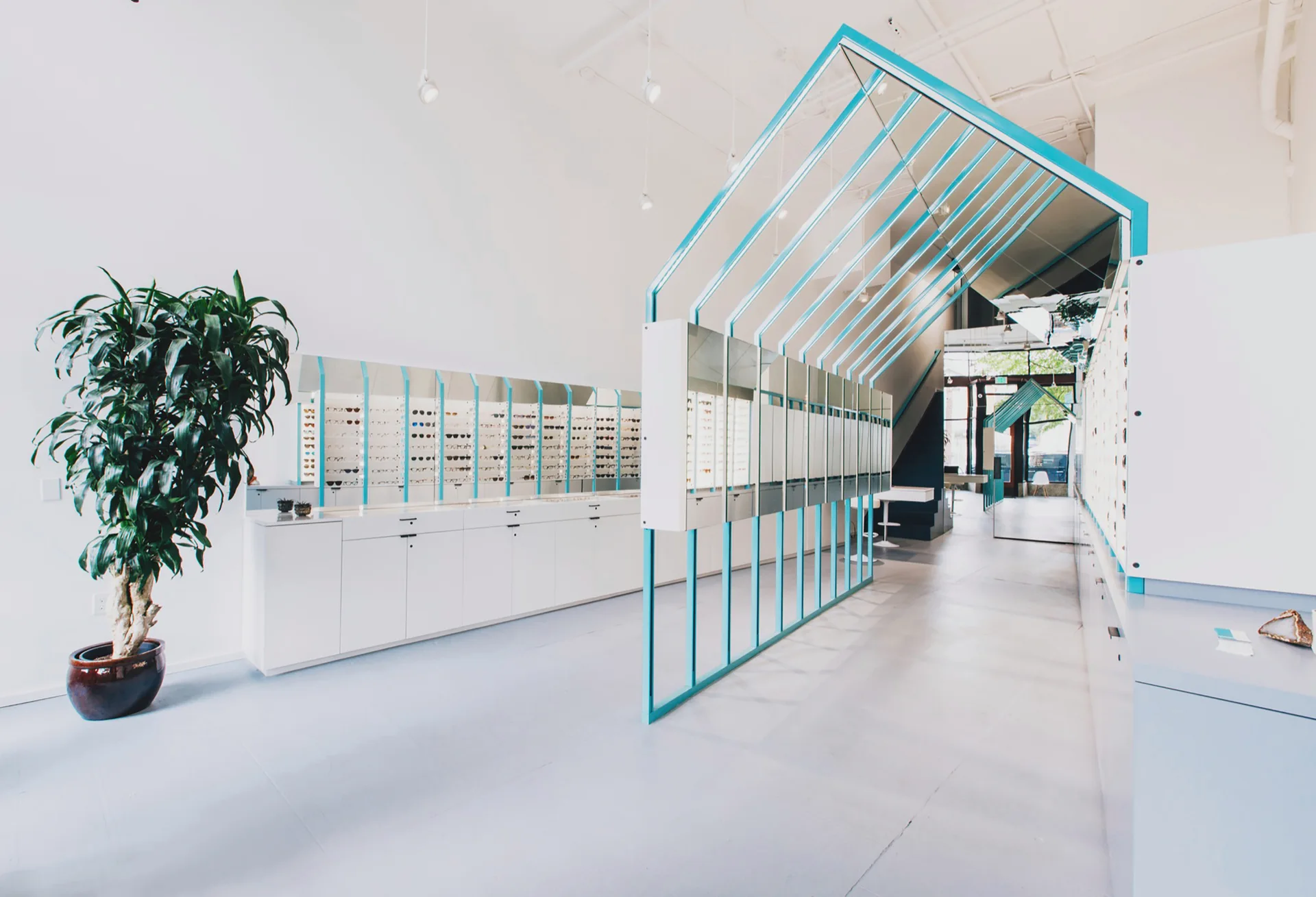 A minimalist retail eyewear store features a large teal metal house-shaped frame, white storage cabinets, and wall-mounted glasses displays.