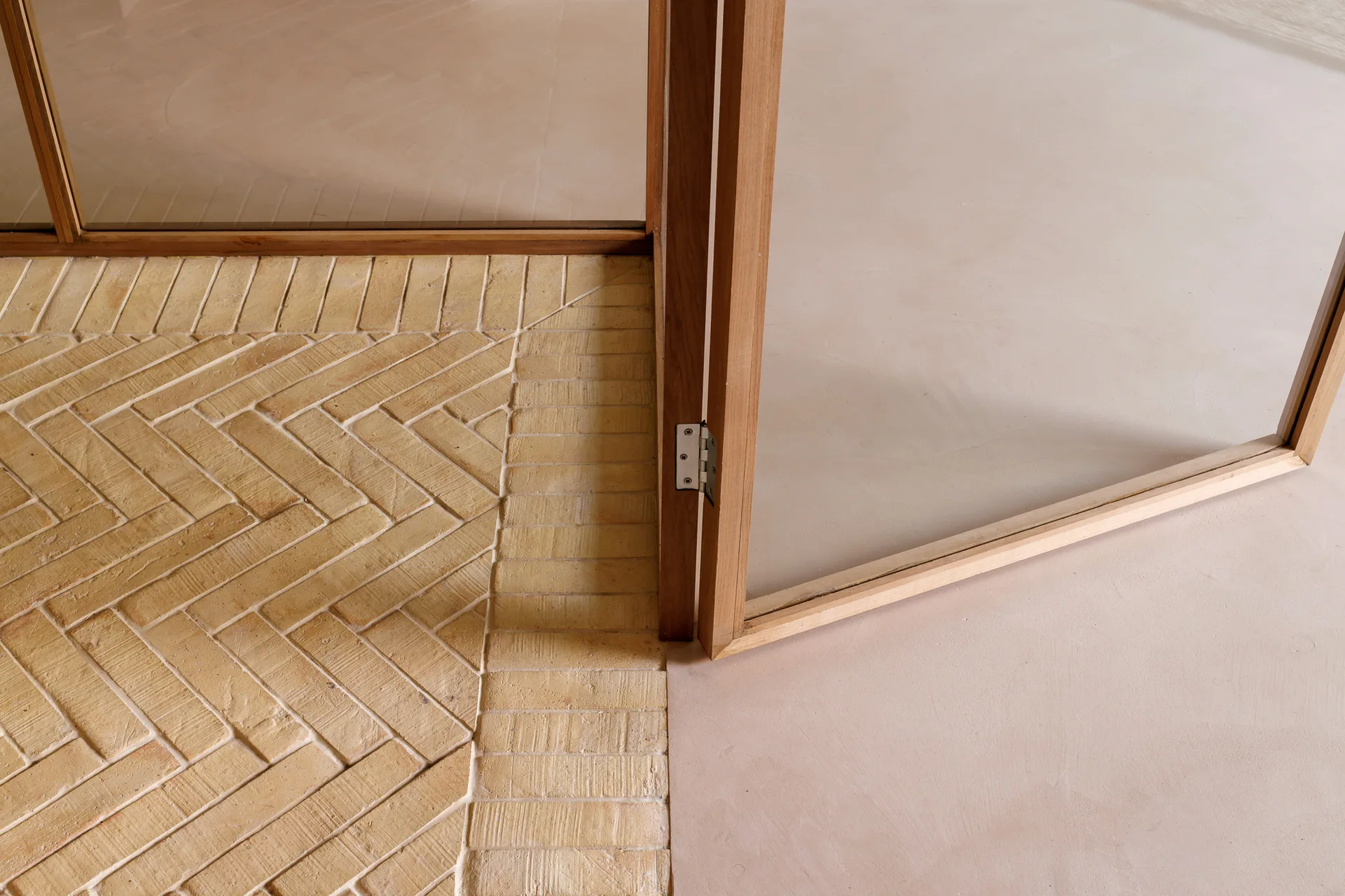 A close-up view of a natural wood door frame where a herringbone pattern yellow brick floor transitions to a smooth beige concrete surface.