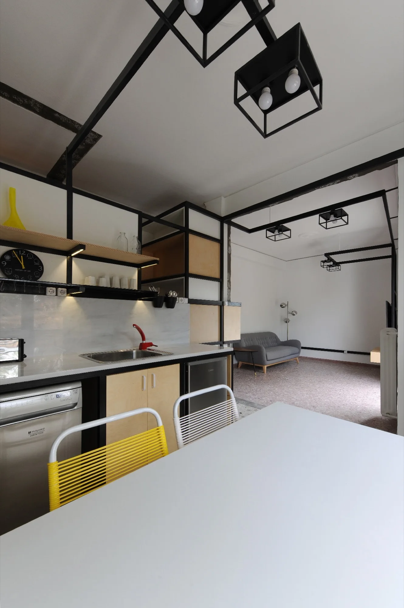 An open-plan kitchen with black geometric ceiling frames, light wood cabinetry, a white dining table, and a yellow wire chair.