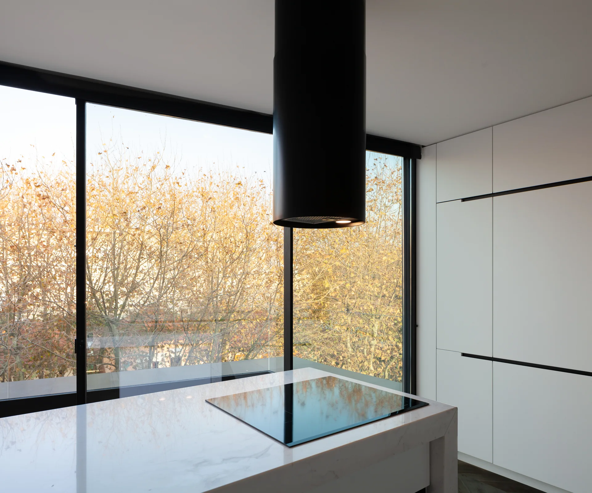 A minimalist kitchen features a white marble island with an integrated cooktop, a black cylindrical range hood, and large floor-to-ceiling windows.