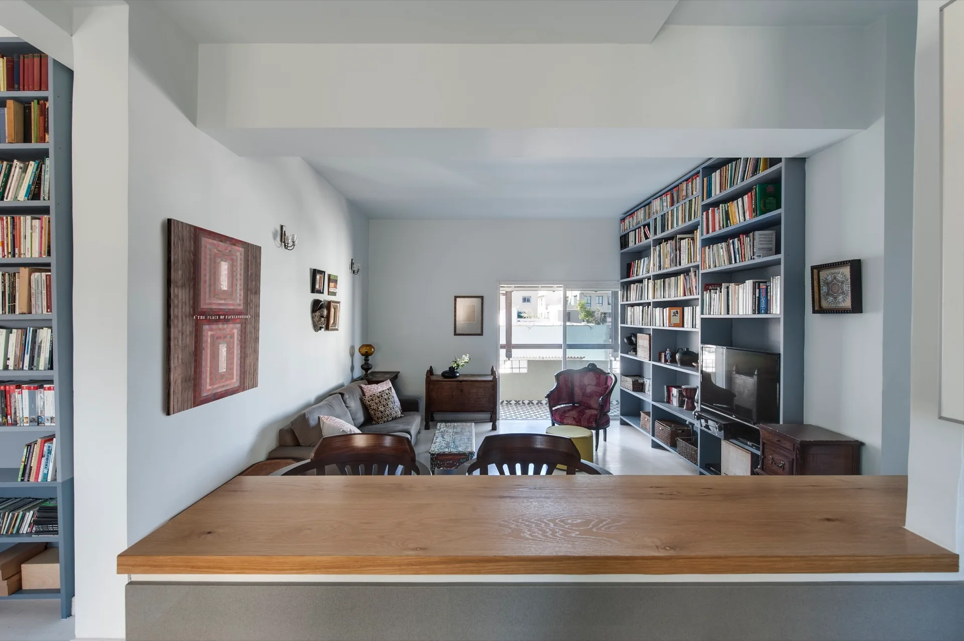 A living room seen from a wooden kitchen counter, featuring a large dusty blue built-in bookshelf, a grey sofa, and an eclectic mix of furniture.