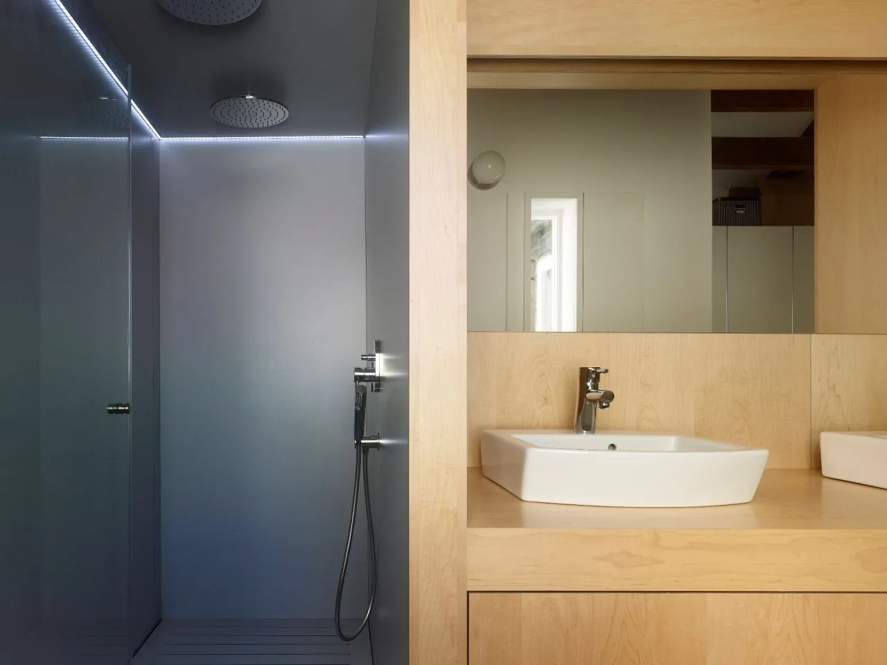 A modern bathroom featuring a light wood vanity with a white ceramic sink adjacent to a dark grey shower stall illuminated by an LED strip.