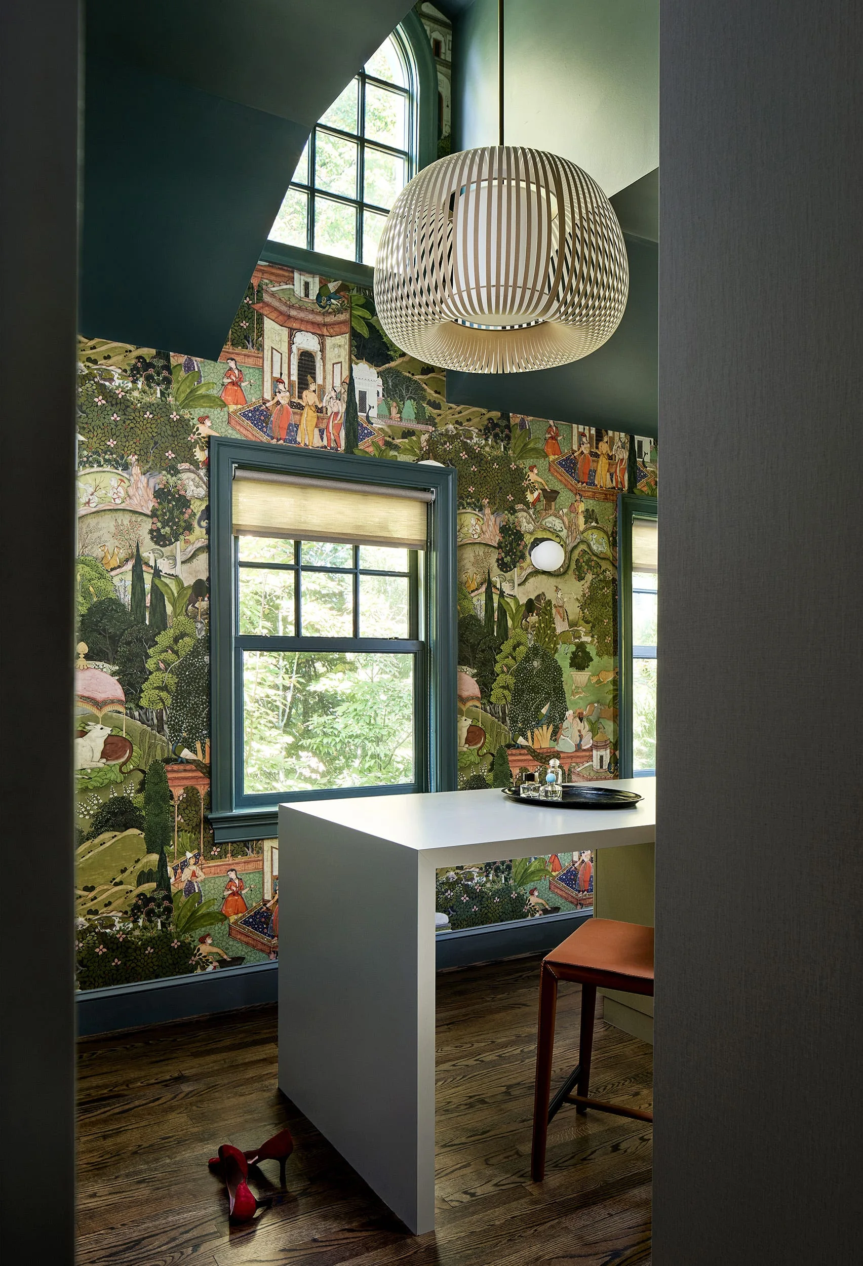 A home office features scenic patterned wallpaper, dark teal painted window trim, a minimalist white desk, and a large slatted pendant light.