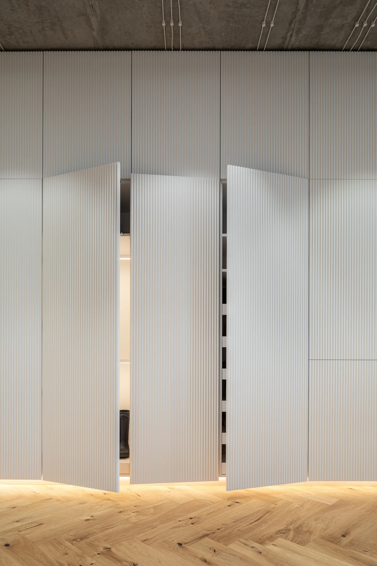 Partially open white fluted closet doors reveal a lit interior above a herringbone hardwood floor and beneath a raw concrete ceiling.