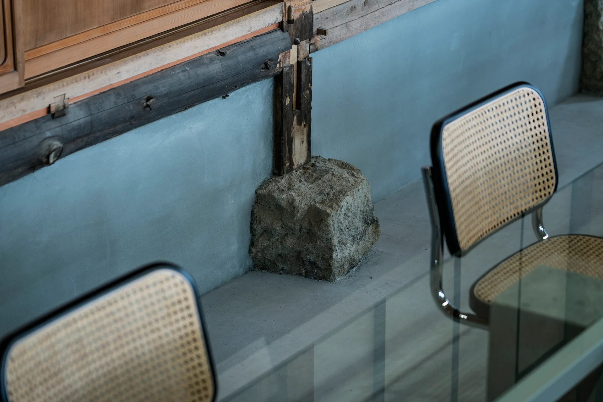 Two woven cane back chairs with chrome frames sit next to a glass table in front of a concrete wall with an exposed timber post and raw stone base.