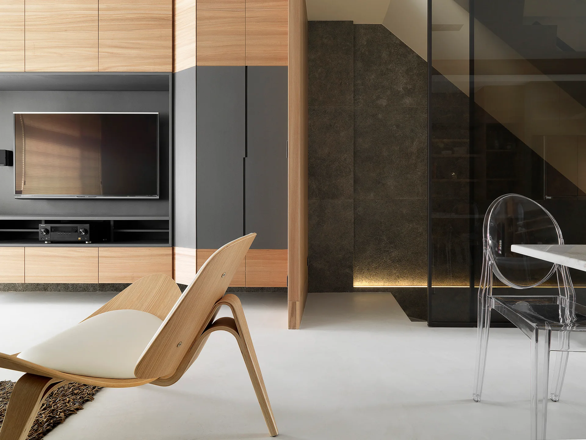 A minimalist living room features a wood and grey built-in media cabinet, a curved plywood armchair, and a transparent dining chair.