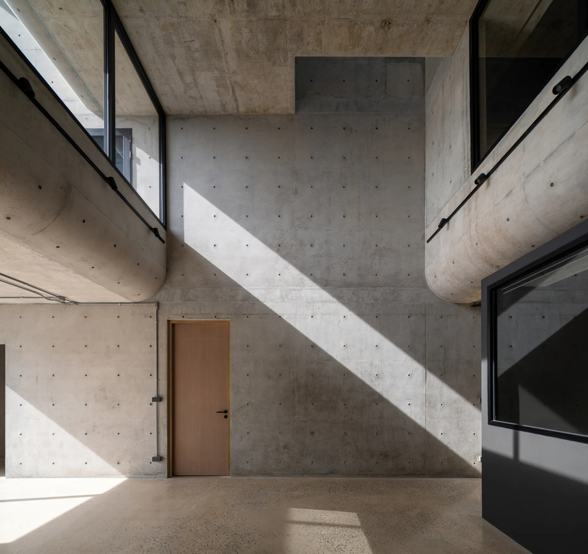A minimalist interior featuring raw concrete walls with form tie holes, a simple wood door, and sunlight streaming through upper black-framed windows.