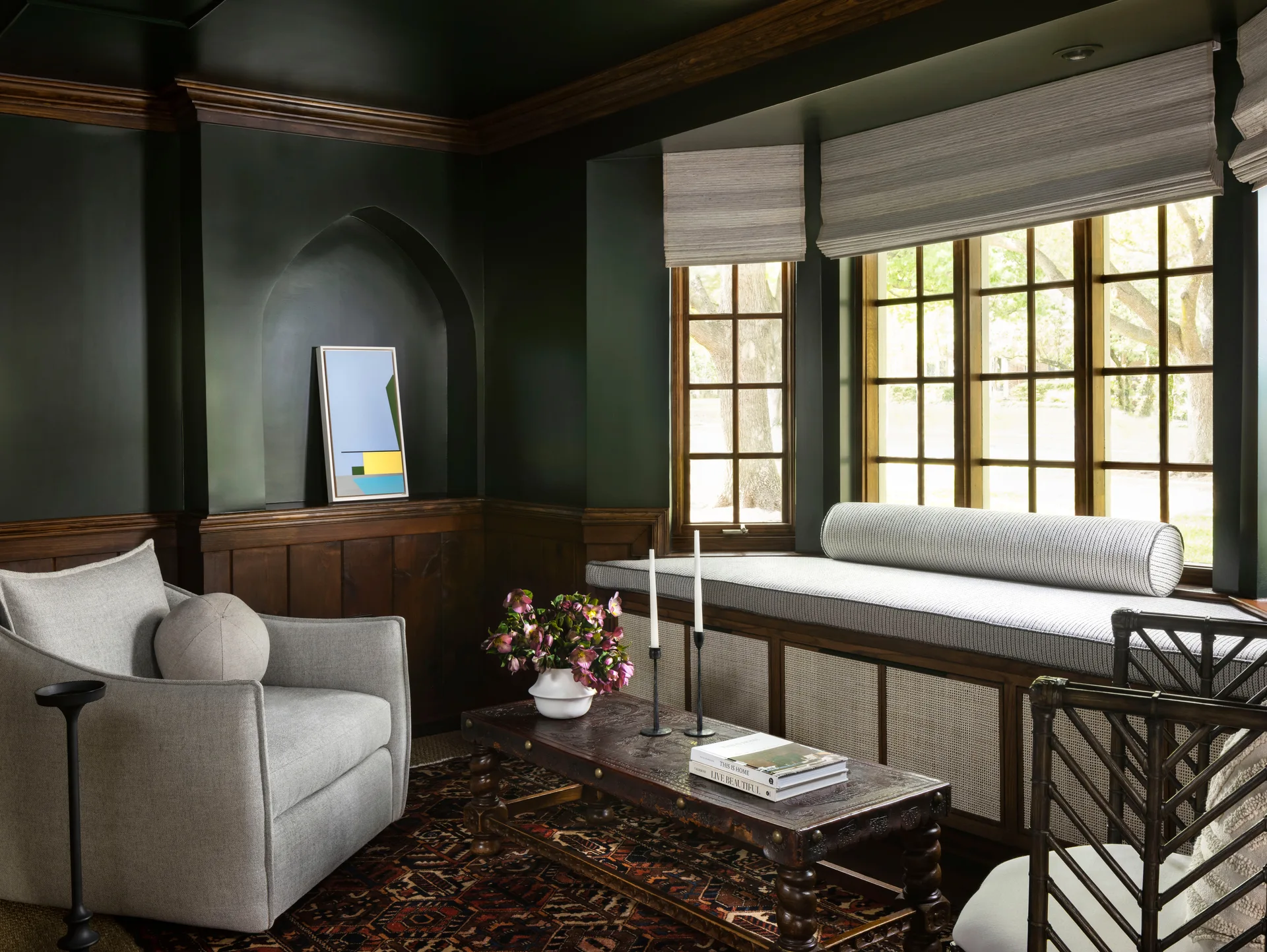 A moody green sitting room features dark wood wainscoting, an arched wall niche with art, an upholstered armchair, and a built-in window bench.