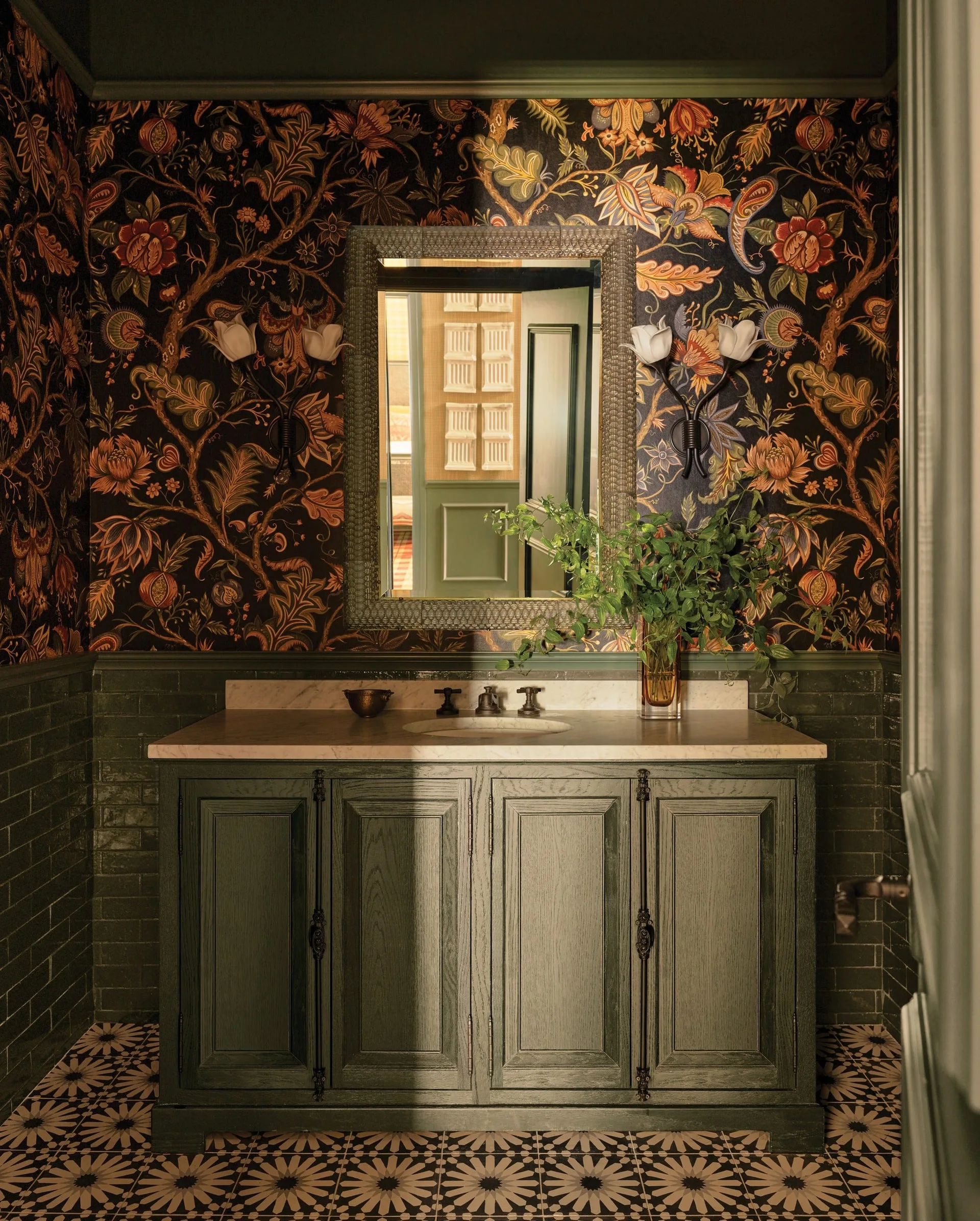 A dark green powder room features botanical wallpaper above tiled wainscoting, a wooden vanity with a marble top, and patterned floor tiles.