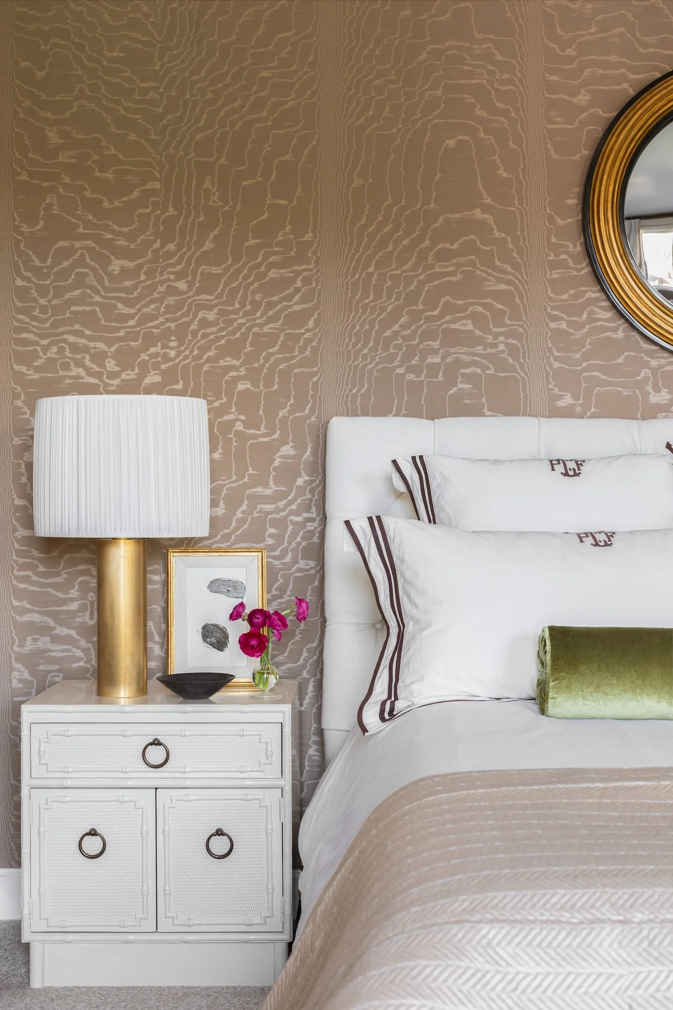 A close-up of a white upholstered bed with monogrammed pillows next to a white bamboo-style nightstand with a brass lamp against patterned wallpaper.