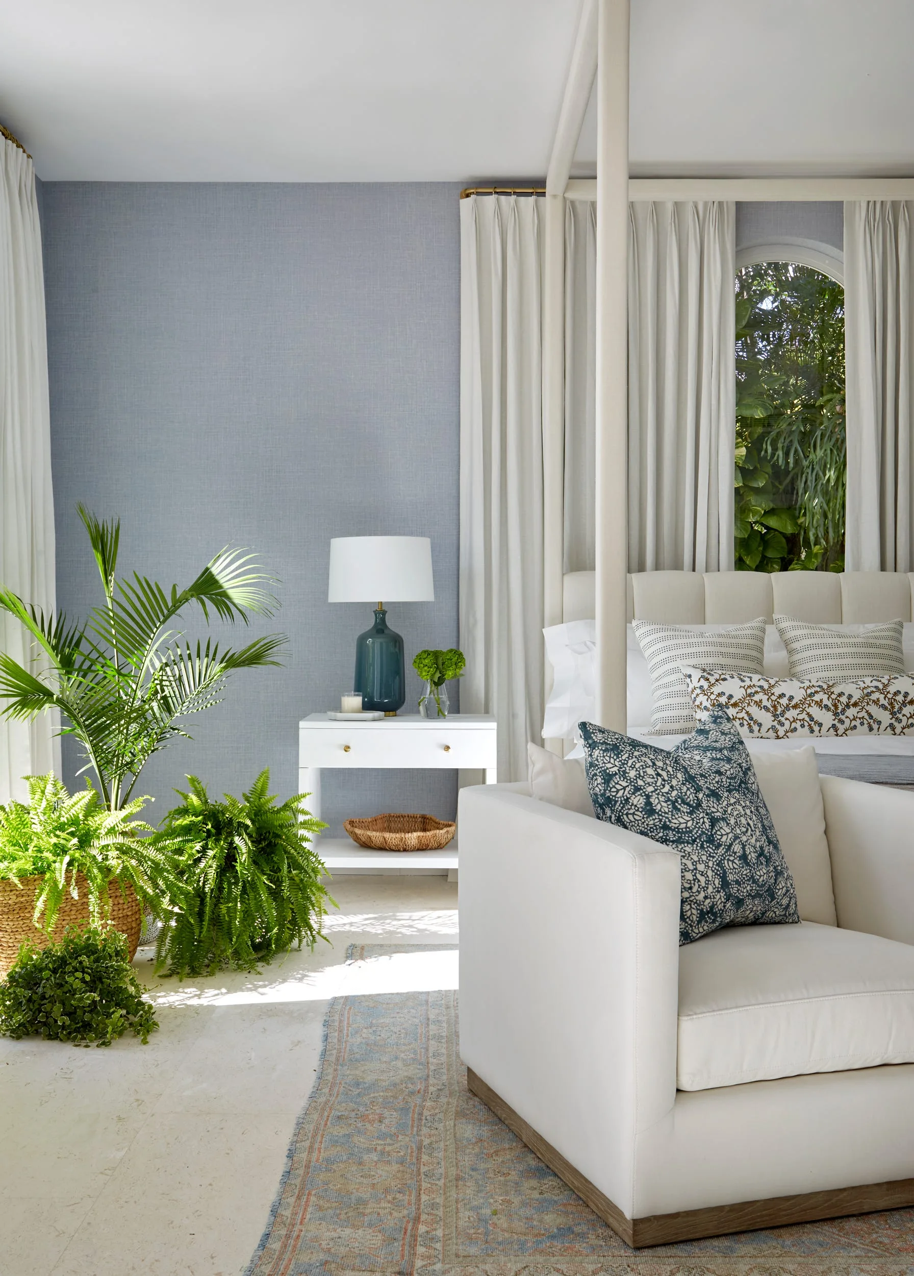 A sunlit bedroom features blue textured wallpaper, a white canopy bed with drapes, a white armchair, and potted green ferns on a stone floor.