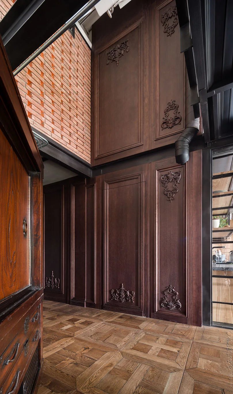 Tall dark brown wood wall paneling with ornate carvings stands opposite a parquet floor, below an exposed red brick wall and black metal structural beam.
